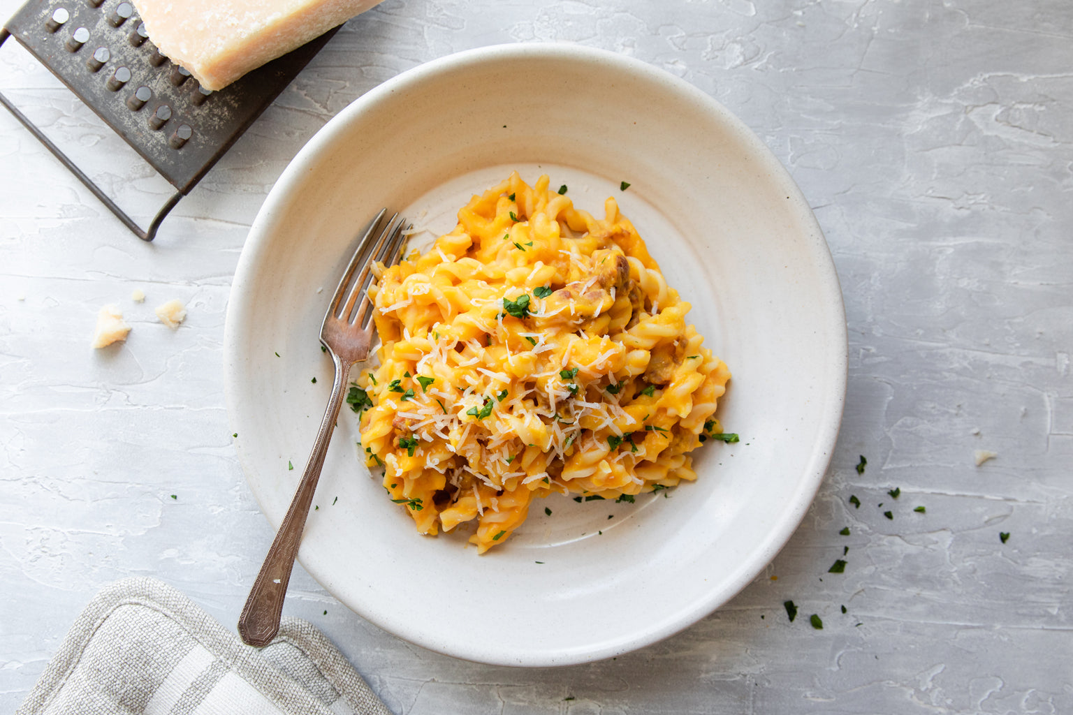 Pumpkin and sausage pasta served in a white bowl with grated cheese and parsley on a gray textured surface