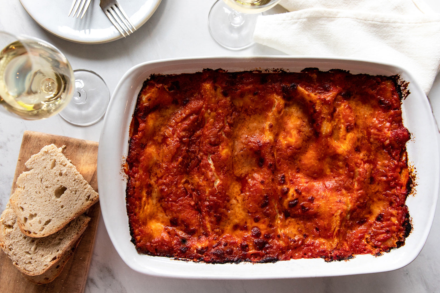 Baked meatless lasagna with browned top in a white dish, on a marble surface with sliced bread and a glass of white wine