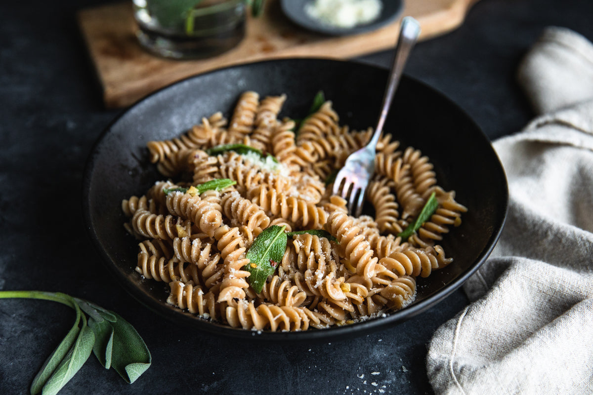 Bowl of fusilli aglio-olio with crispy sage and grated cheese on a dark plate, fork resting in the pasta