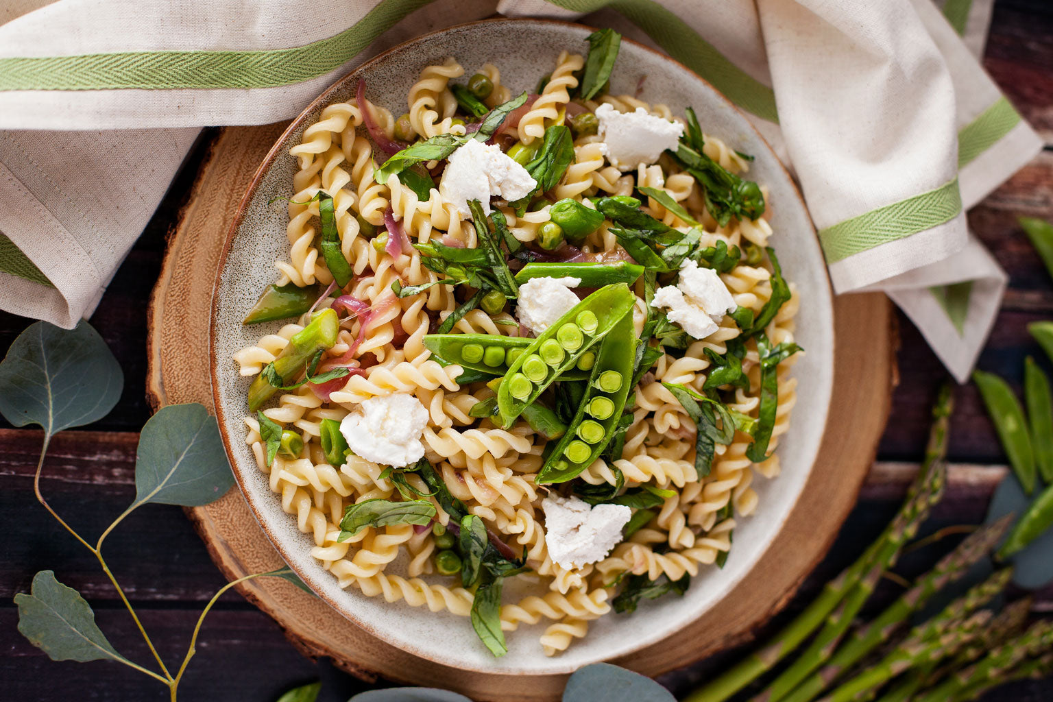 Bowl of fusilli pasta tossed with spring vegetables like snap peas, asparagus, and peas, topped with dollops of ricotta on a wooden board with a striped napkin