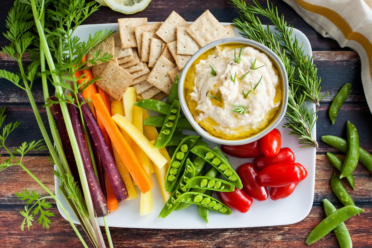 White bean and rosemary spread in a small bowl with olive oil, served on a platter with crackers, snap peas, cherry tomatoes, and carrot sticks
