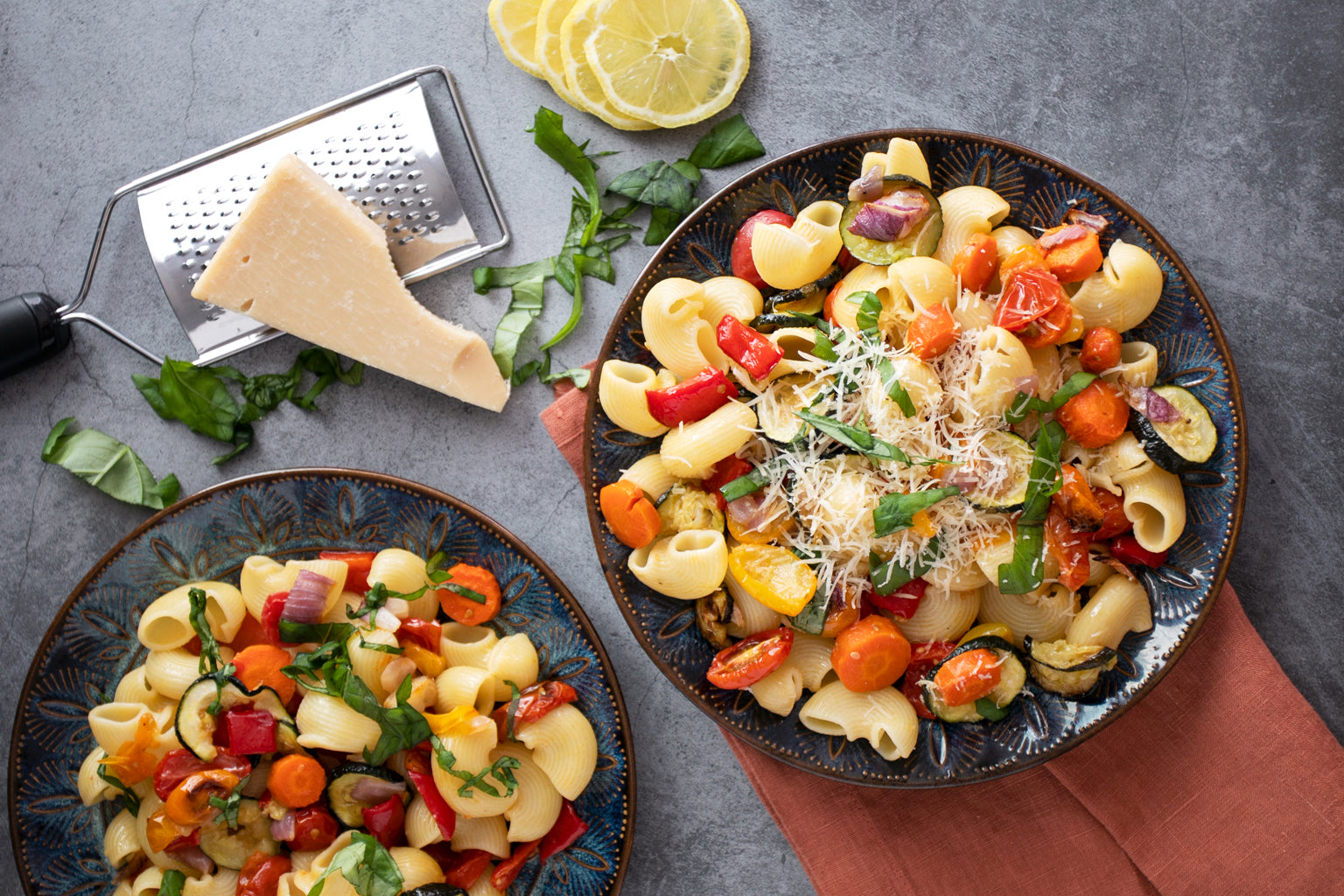 Two bowls of roasted summer vegetable pasta salad with cherry tomatoes, zucchini, carrots and grated cheese on a gray surface