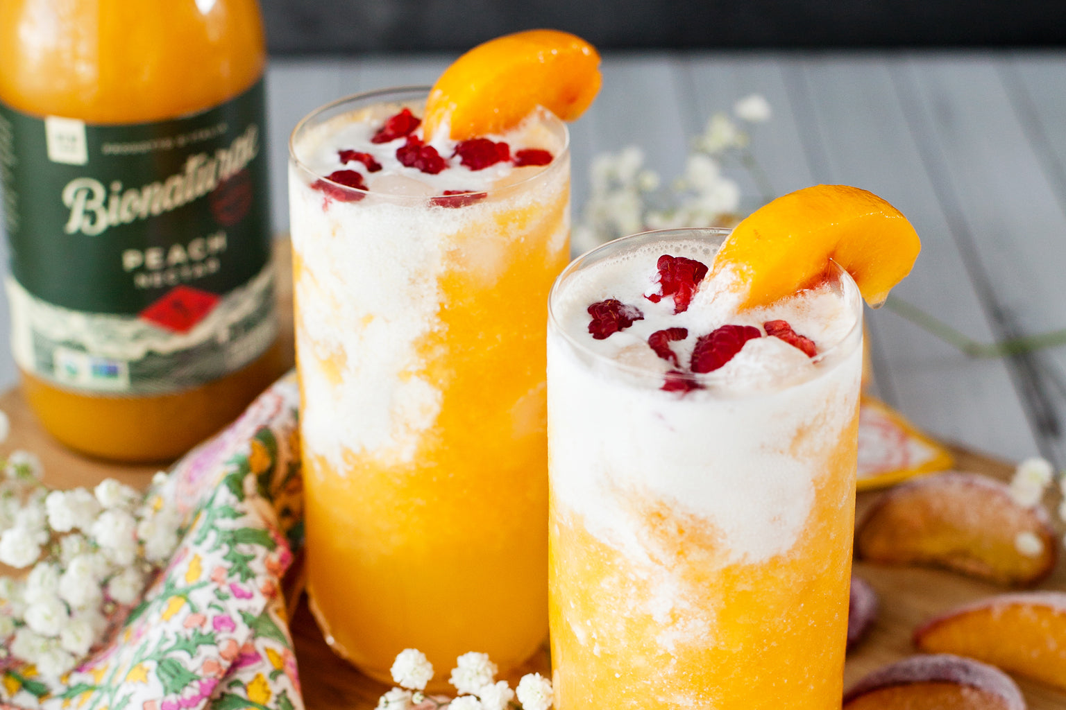 Two peach Italian sodas with frothy cream and raspberries, garnished with peach slices on a wooden board with a Bionaturae bottle in the background