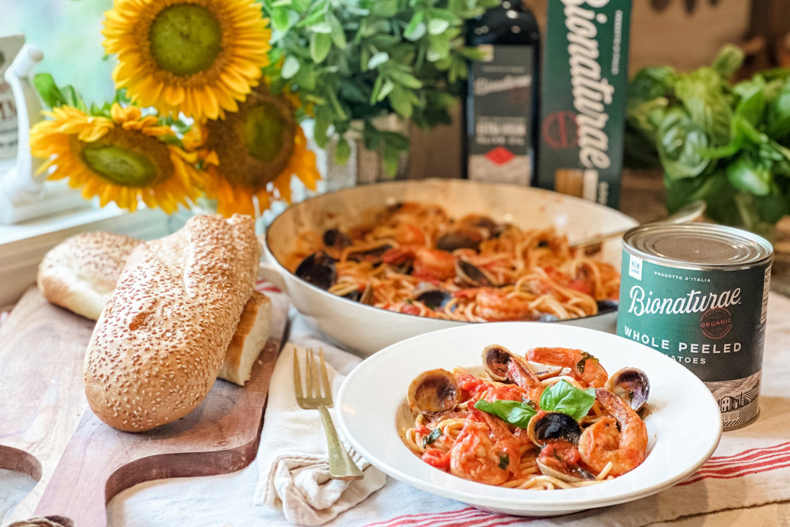 Bowl of spaghetti with clams and shrimp in tomato sauce on a table with a skillet of pasta, sesame bread, sunflowers, and a can of Bionaturae whole peeled tomatoes