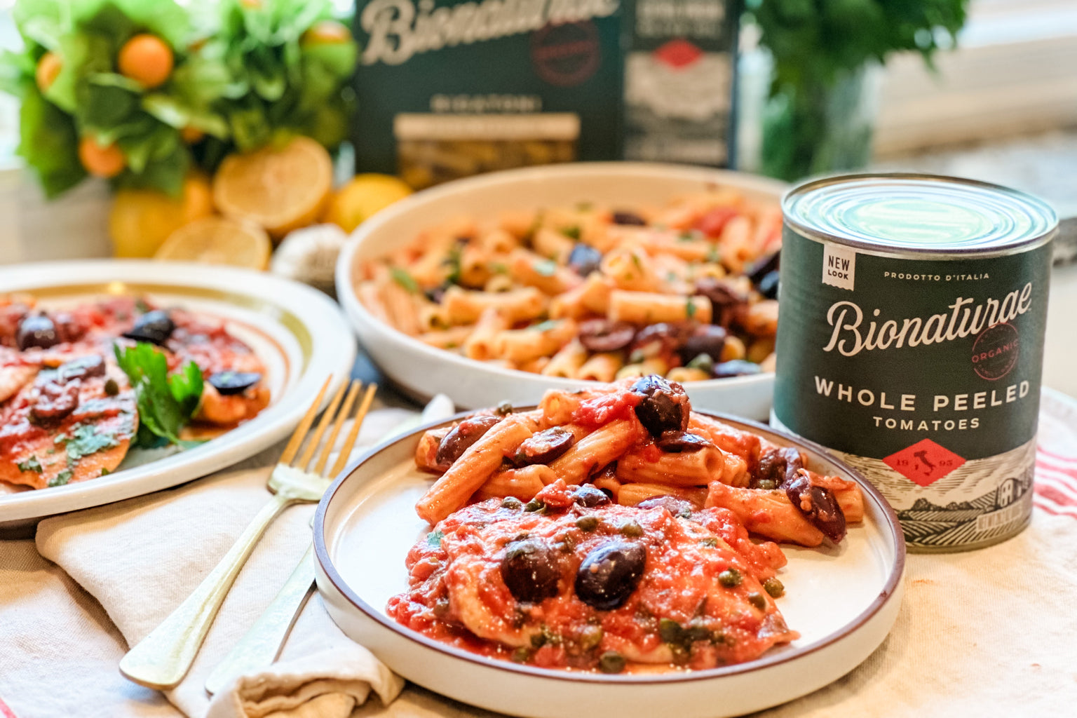 Plated chicken in white wine, caper and tomato sauce with penne and olives on a kitchen table, a can of tomatoes and lemons in the background