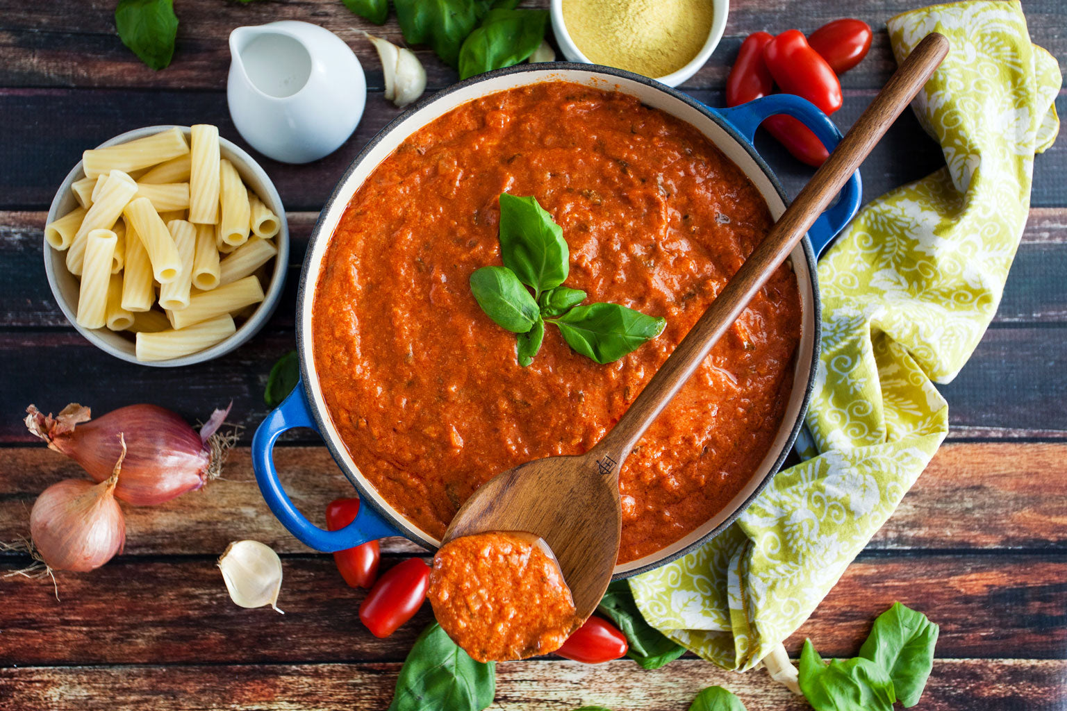 Tomato basil cream sauce in a blue pot with a wooden spoon, basil garnish, uncooked pasta and shallots on a rustic wooden table