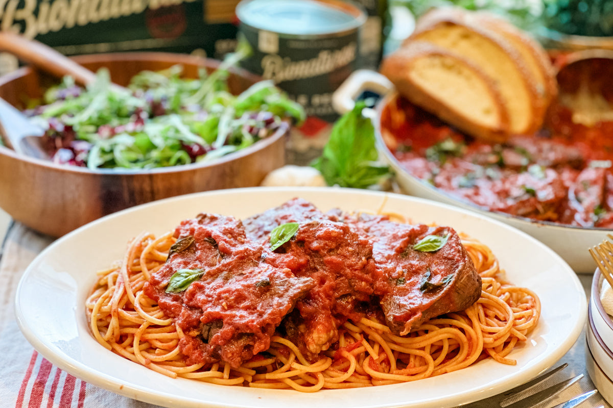 Steak pizzaiola on spaghetti with tomato sauce and basil, white plate with salad and bread in the background