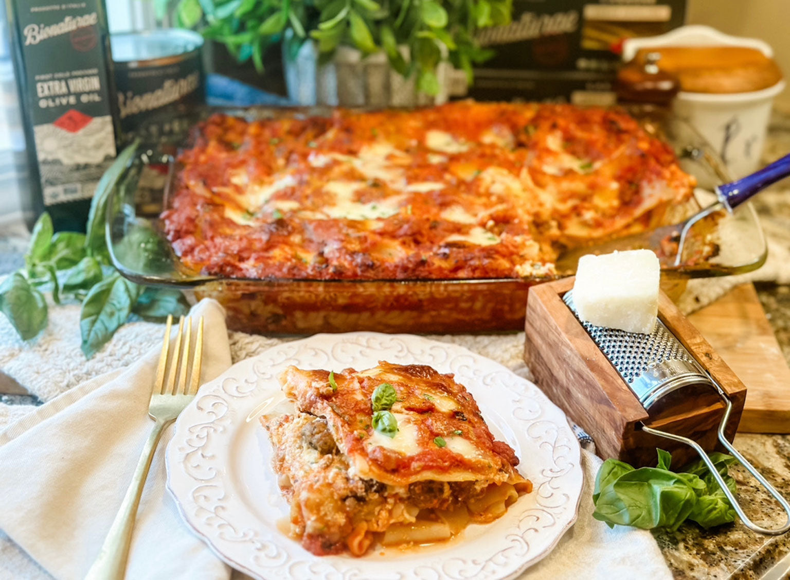 Neapolitan-style lasagna slice on a white plate with fresh basil, with a glass baking dish of lasagna and a cheese grater on a kitchen counter