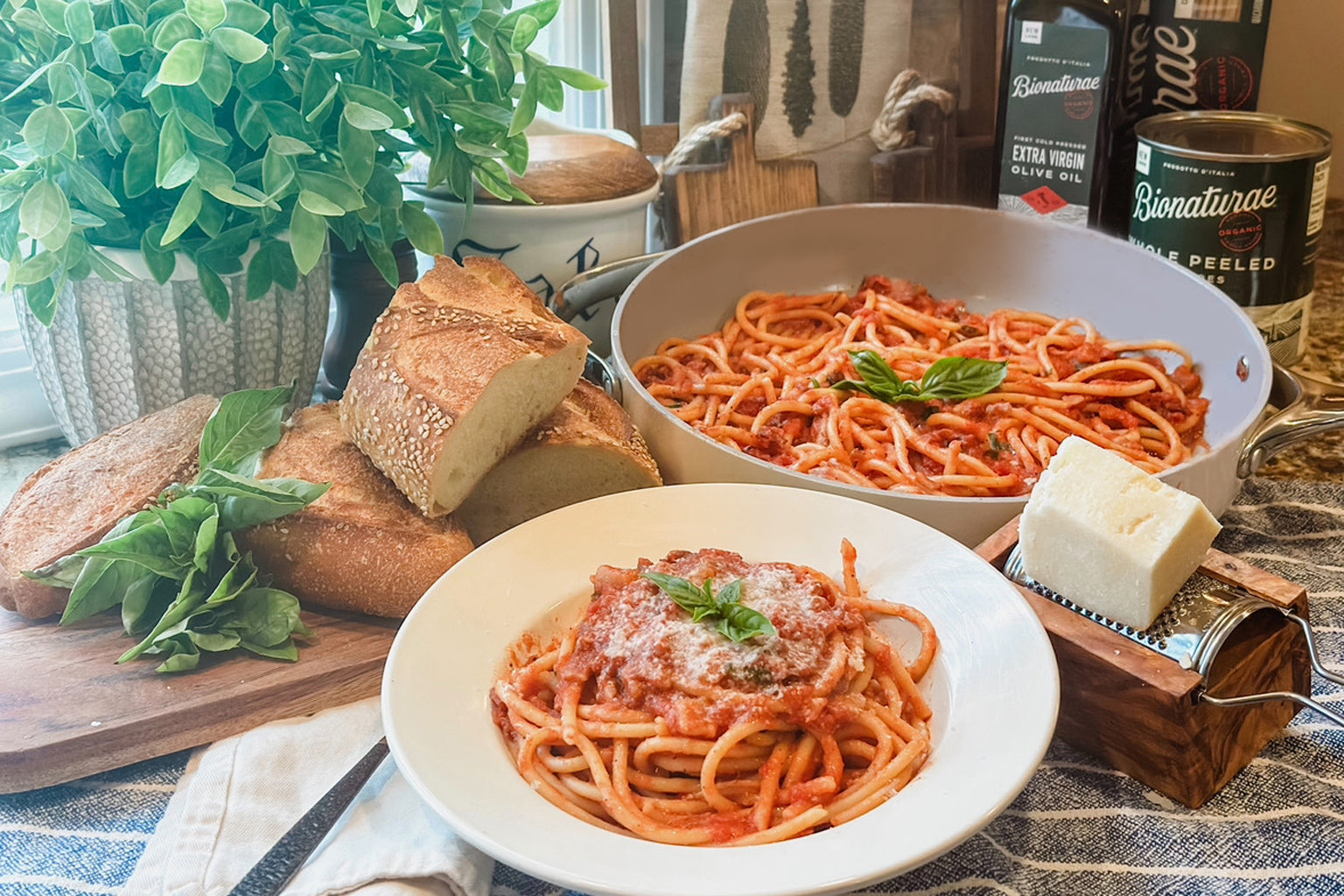 Bucatini amatriciana served in a white bowl topped with grated cheese, with crusty bread, fresh basil, and a pan of pasta on a kitchen table