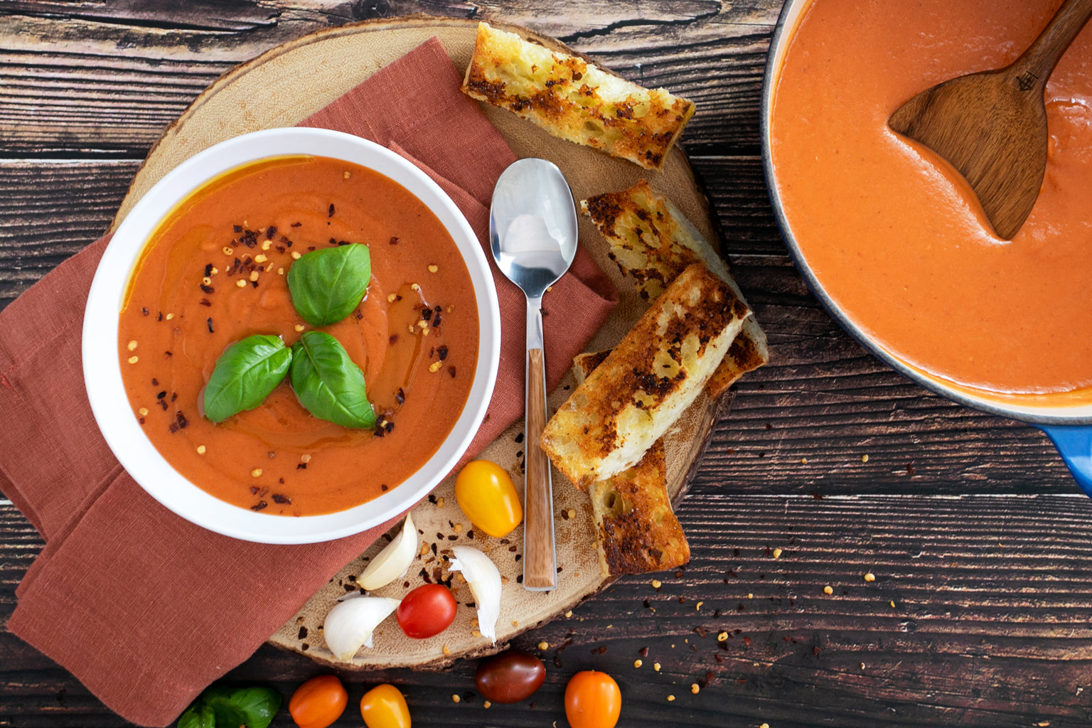 Overhead view of a bowl of tomato soup garnished with basil and red pepper flakes, with toasted bread and a spoon on a wooden board