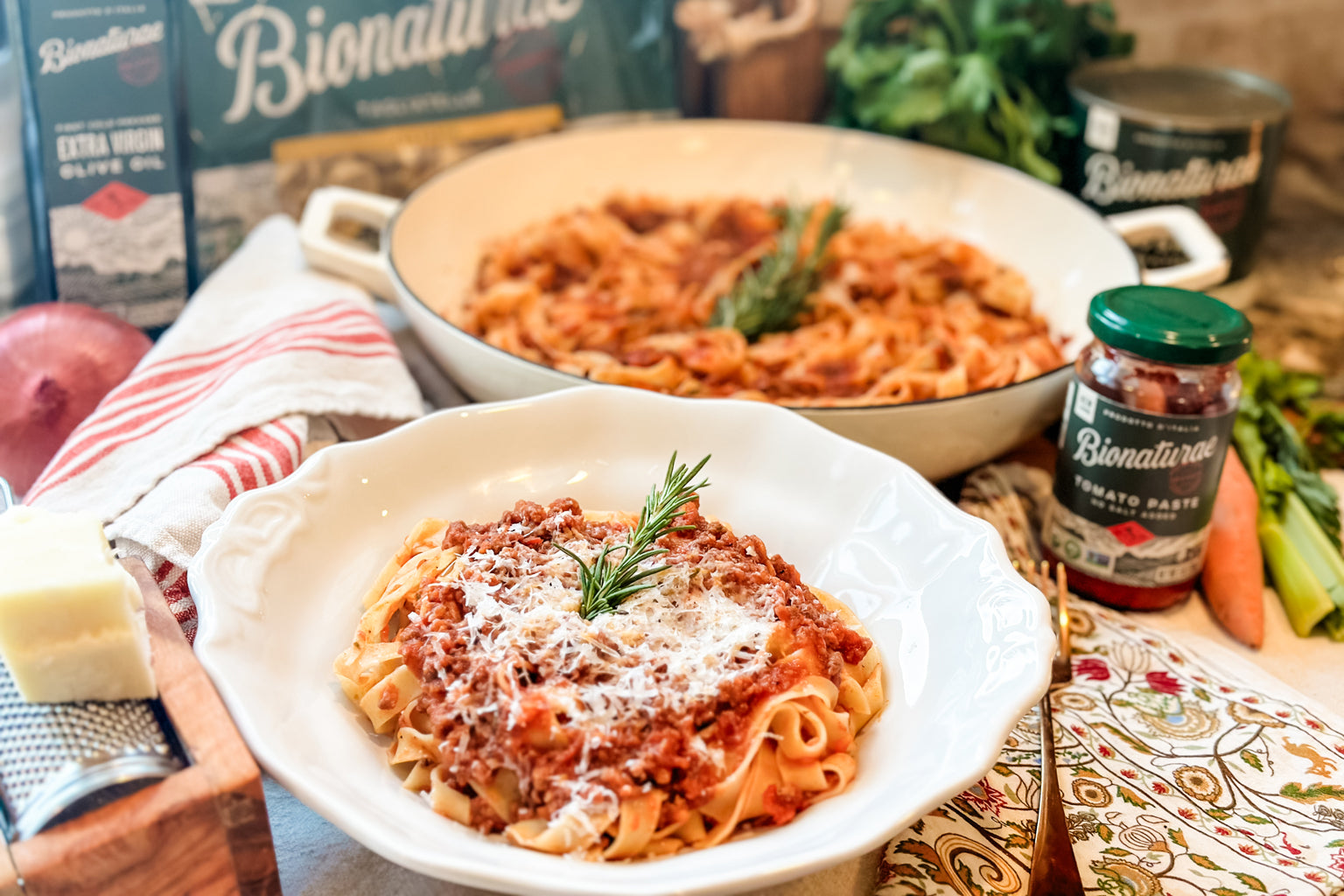 White bowl of tagliatelle topped with Tuscan ragù, grated cheese, and rosemary, with a jar of tomato paste and skillet behind