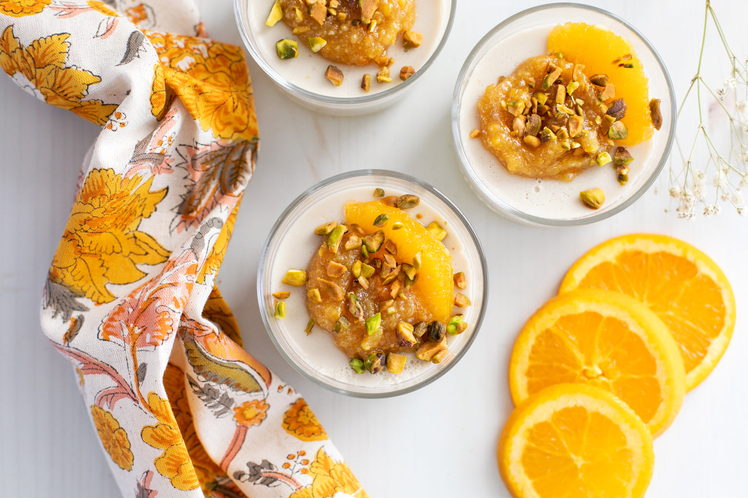 Top-down view of panna cotta in glass jars topped with citrus ginger compote and chopped pistachios, beside orange slices and a floral napkin