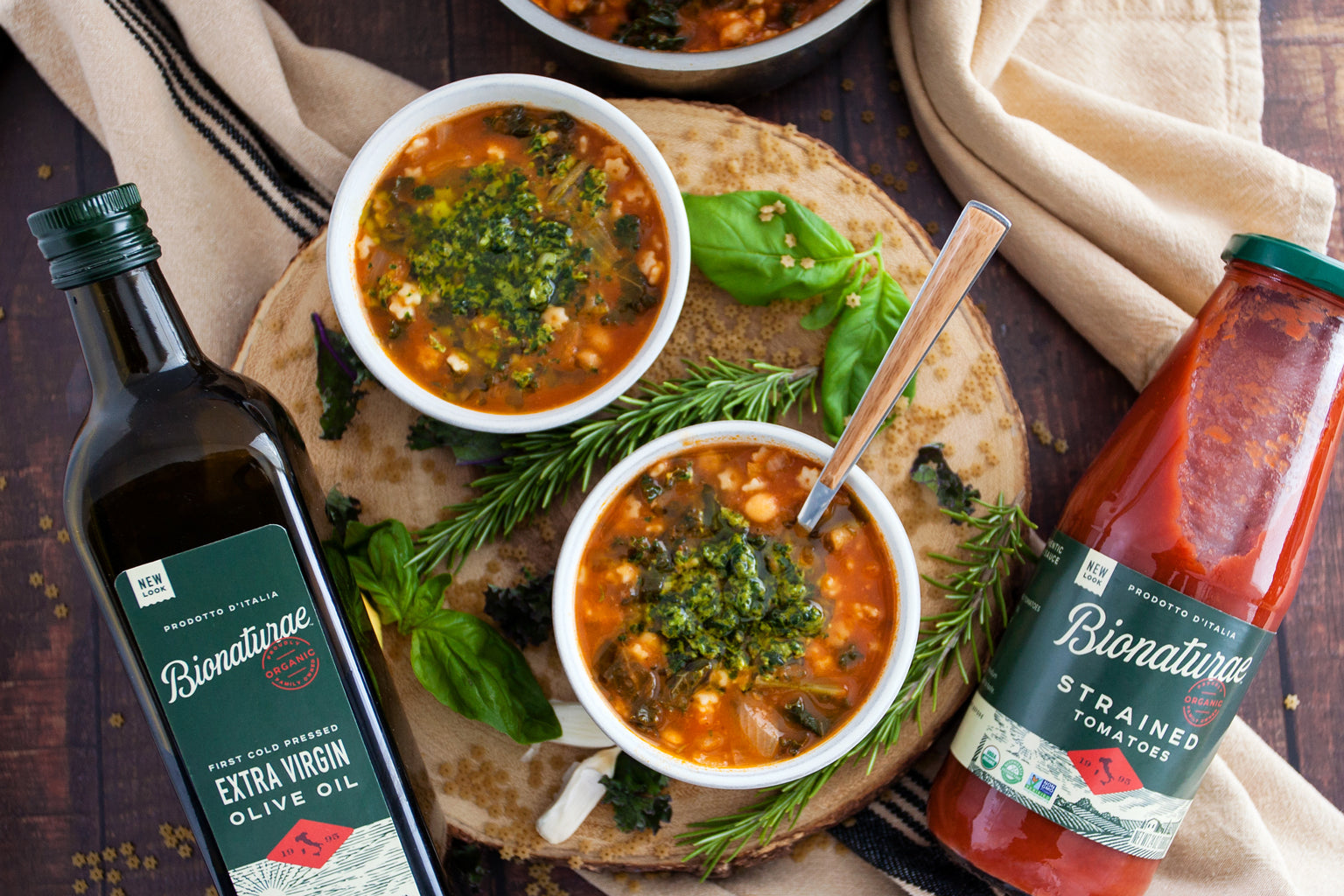 Two bowls of rosemary minestrone topped with spinach pesto on a wooden board with Bionaturae olive oil and strained tomatoes