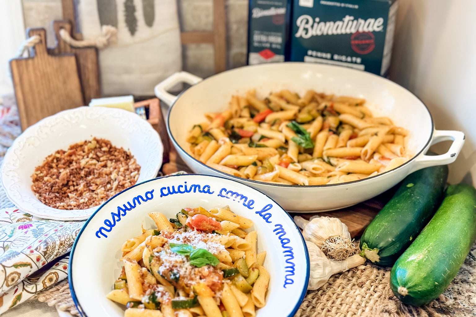 Penne with zucchini, tomatoes, and basil in a bowl beside a skillet of pasta, fresh zucchinis, garlic, and breadcrumbs