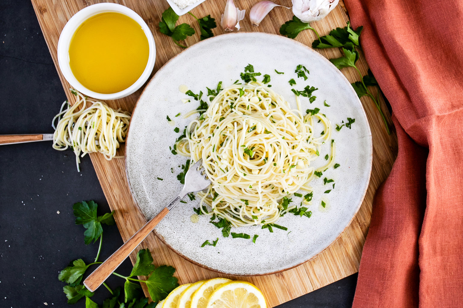 Plate of capellini aglio e olio with parsley, lemon wedge, and a fork twirling pasta on a wooden board beside a bowl of olive oil