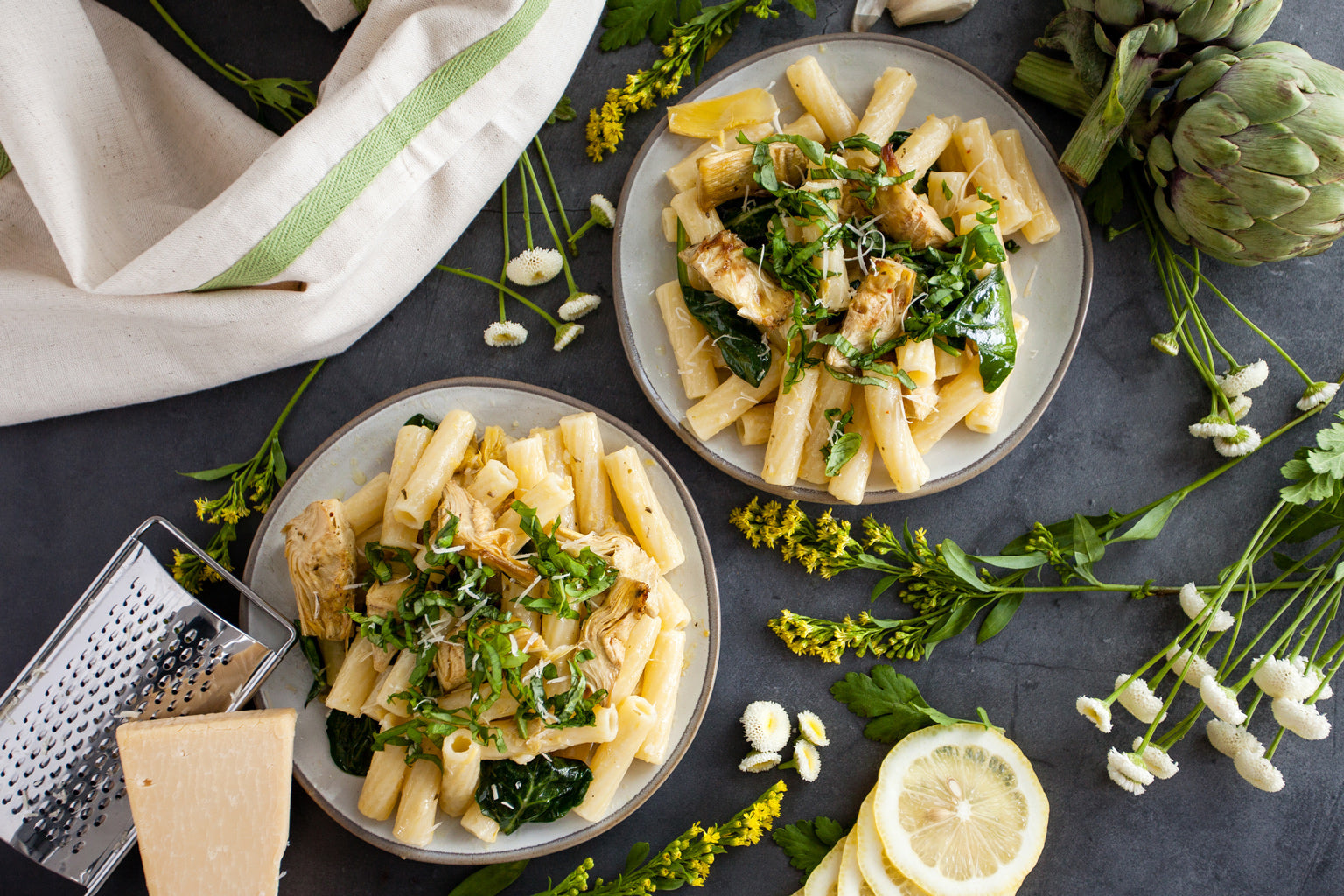Two bowls of lemon basil rigatoni served with roasted artichokes, wilted greens, lemon slices, and a wedge of Parmesan on a dark tabletop