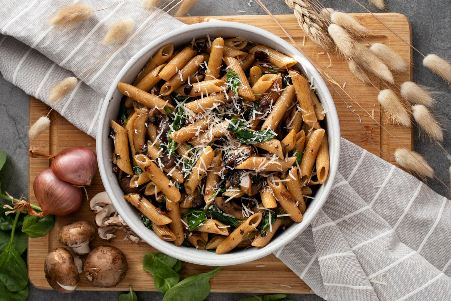 Bowl of whole wheat penne with white wine sauteed mushrooms, garlicky spinach, and grated cheese on a wooden board