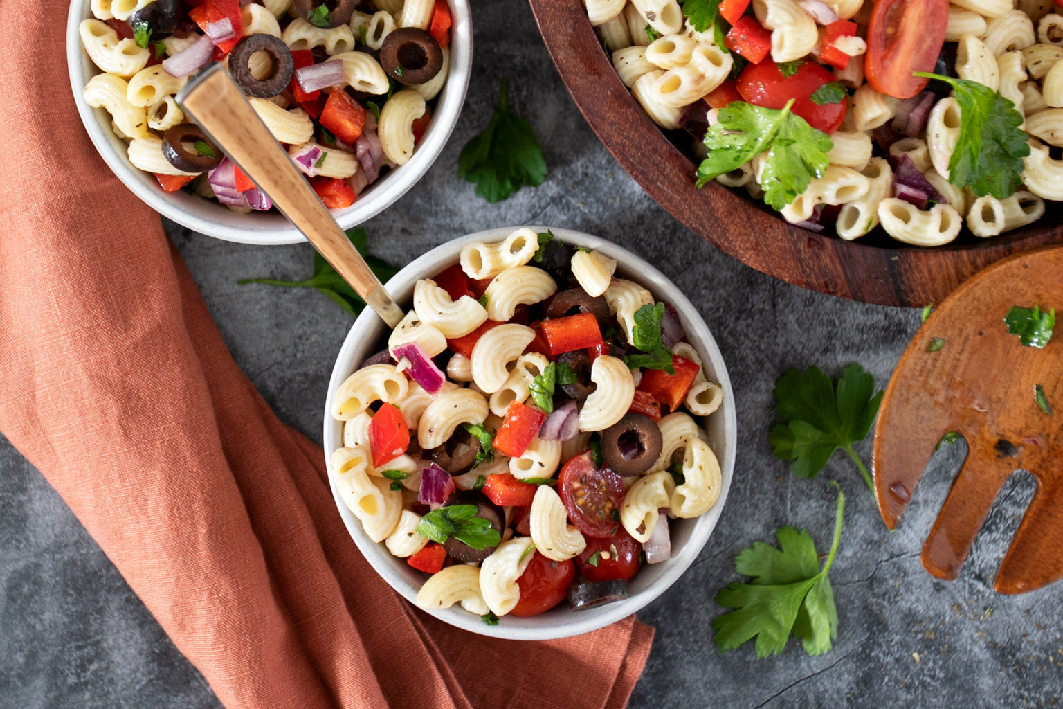 Three bowls of gluten-free Italian pasta salad served with a wooden serving fork and rust-colored napkin on a gray stone surface
