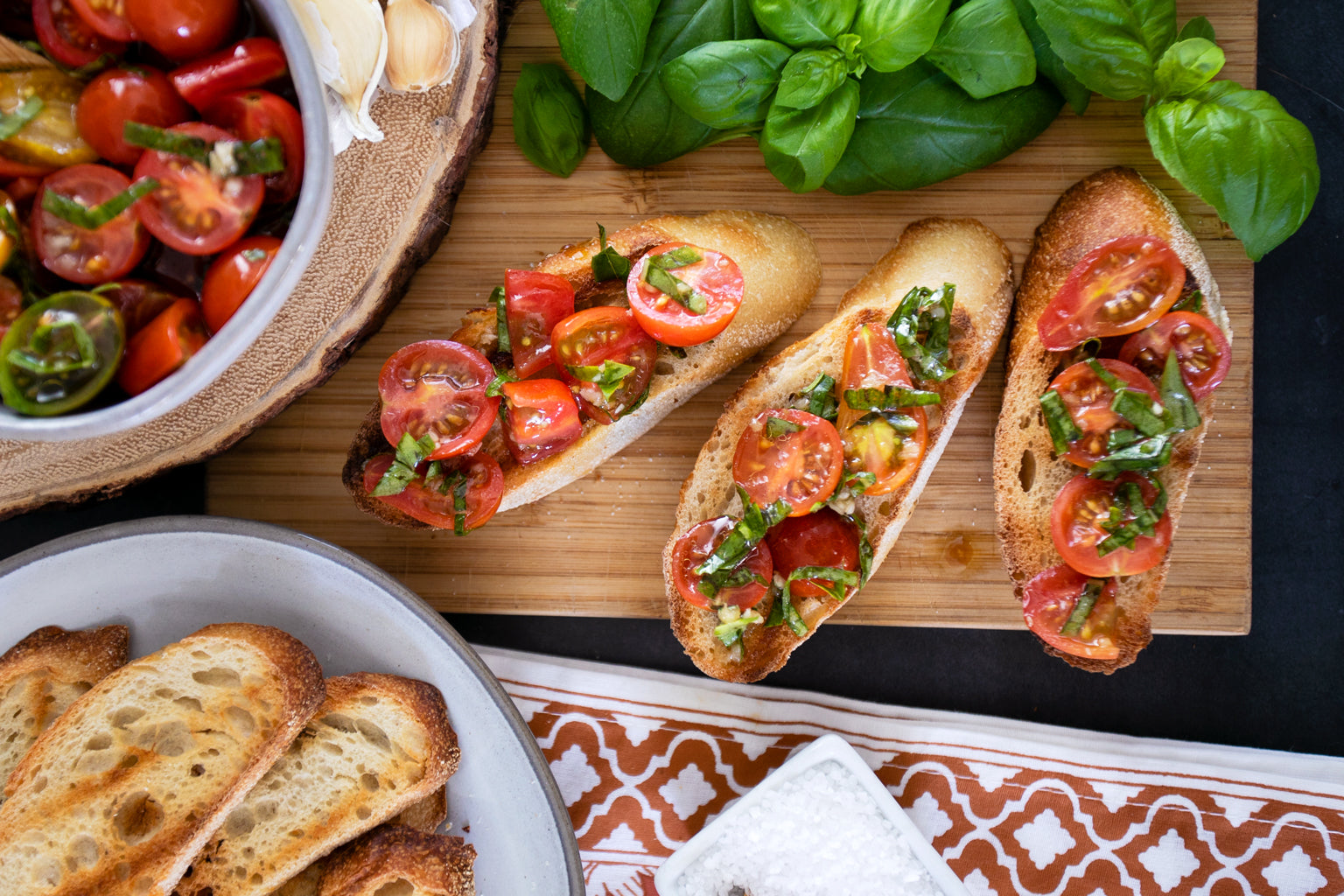 Overhead view of cherry tomato bruschetta on toasted baguette slices with chopped basil and a bowl of tomato salad on a wooden board