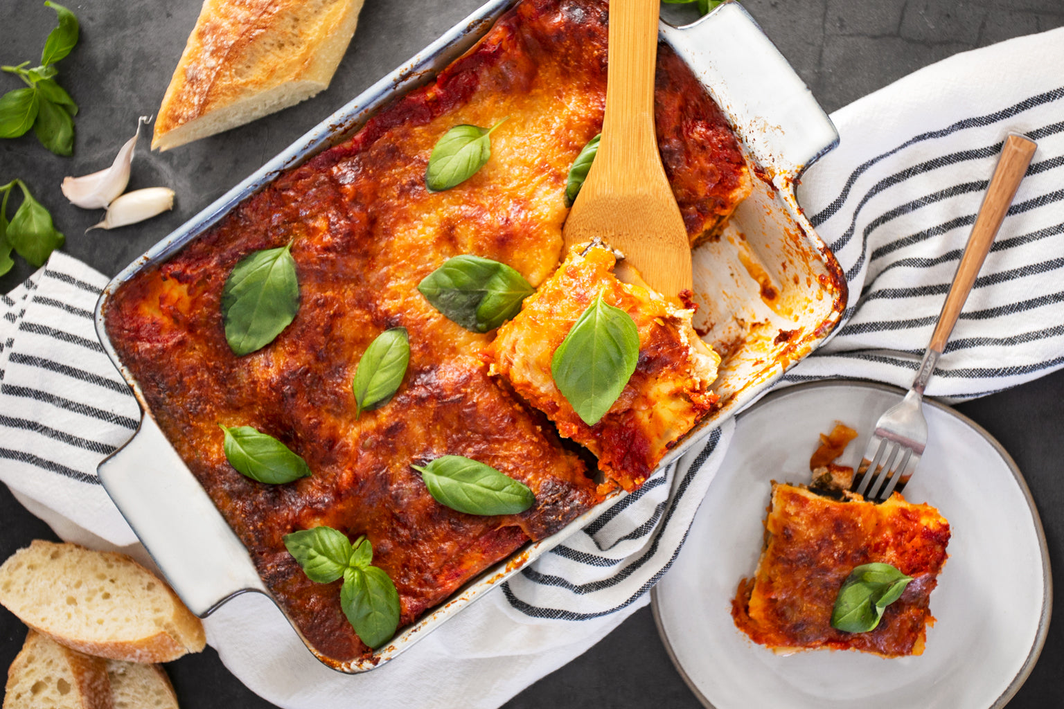 Overhead view of eggplant lasagna with golden-browned cheese and basil, slice served on plate with bread