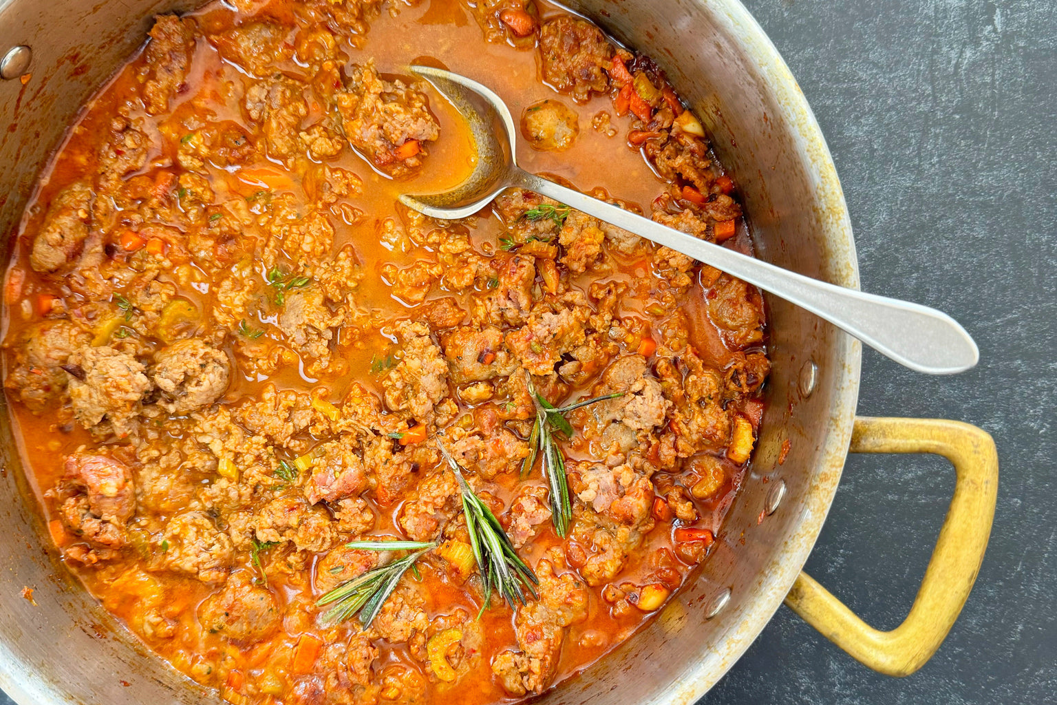 Sausage ragù simmering in a large pot with rosemary sprigs and a serving spoon on a dark countertop