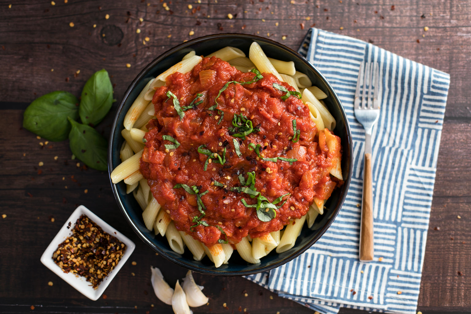 Bowl of penne arrabbiata with spicy tomato sauce and basil garnish on a wooden table with striped napkin and fork