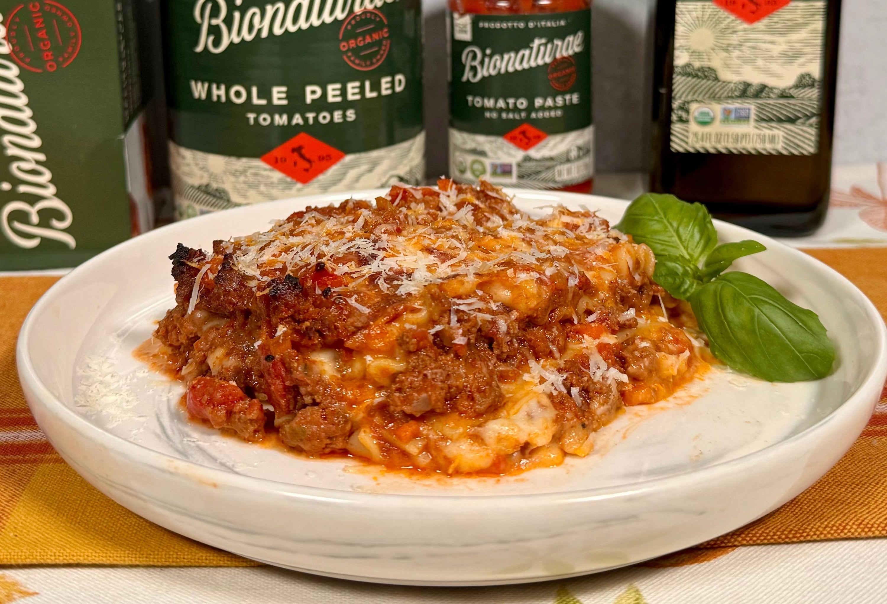 Slice of lasagna alla Bolognese on a white plate with grated cheese and fresh basil, cans and a bottle behind it