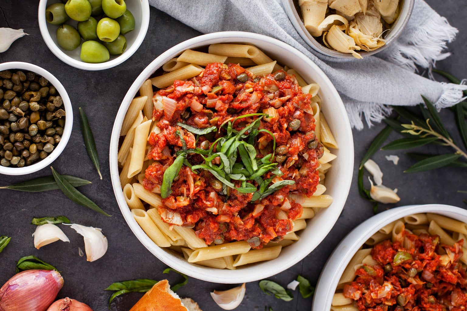 Top-down view of bowls of penne puttanesca topped with basil, with olives, capers, garlic, and shallot nearby