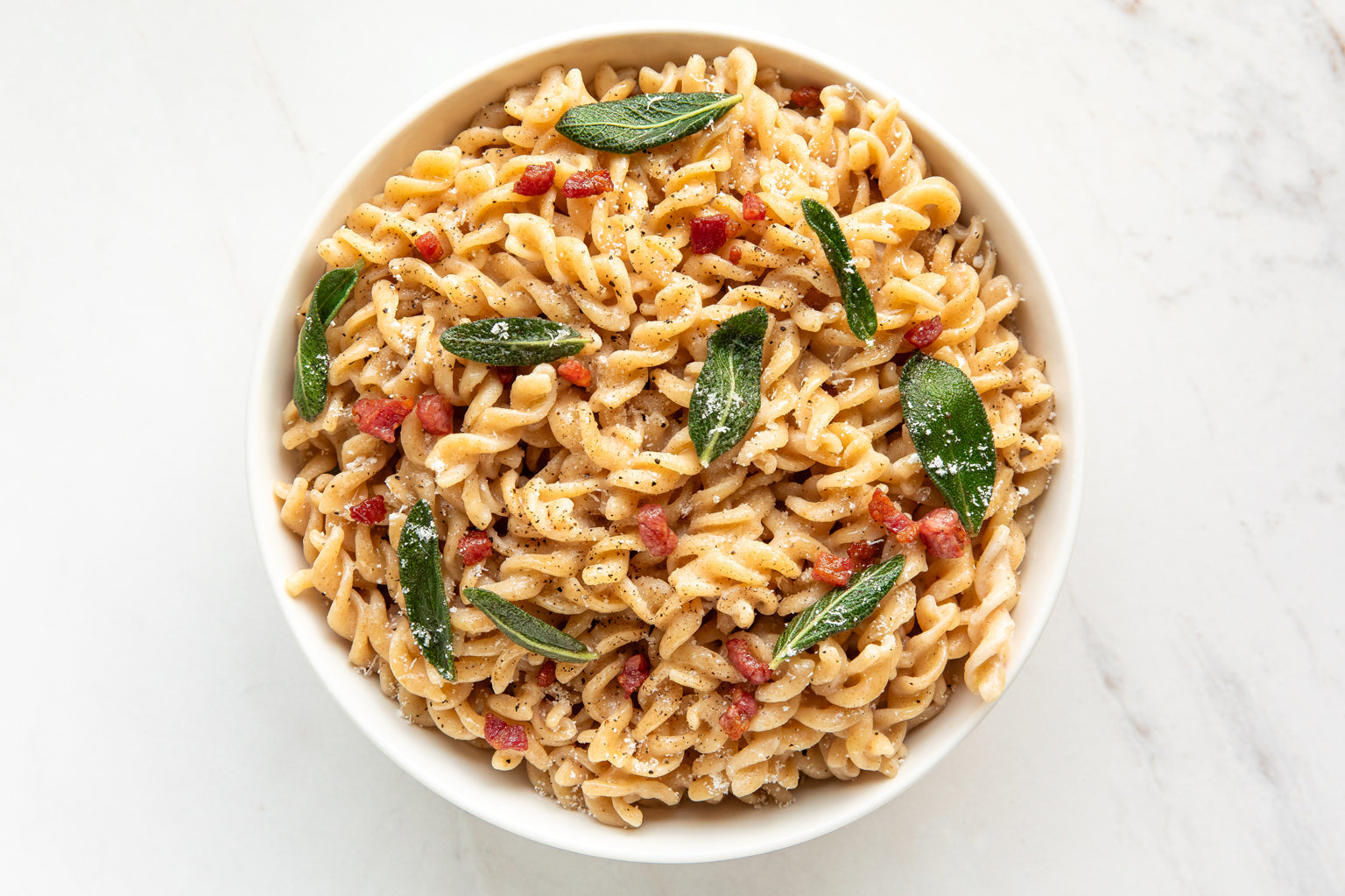 Bowl of fusilli pasta tossed in browned butter with fried sage leaves and crispy pancetta bites on white marble background