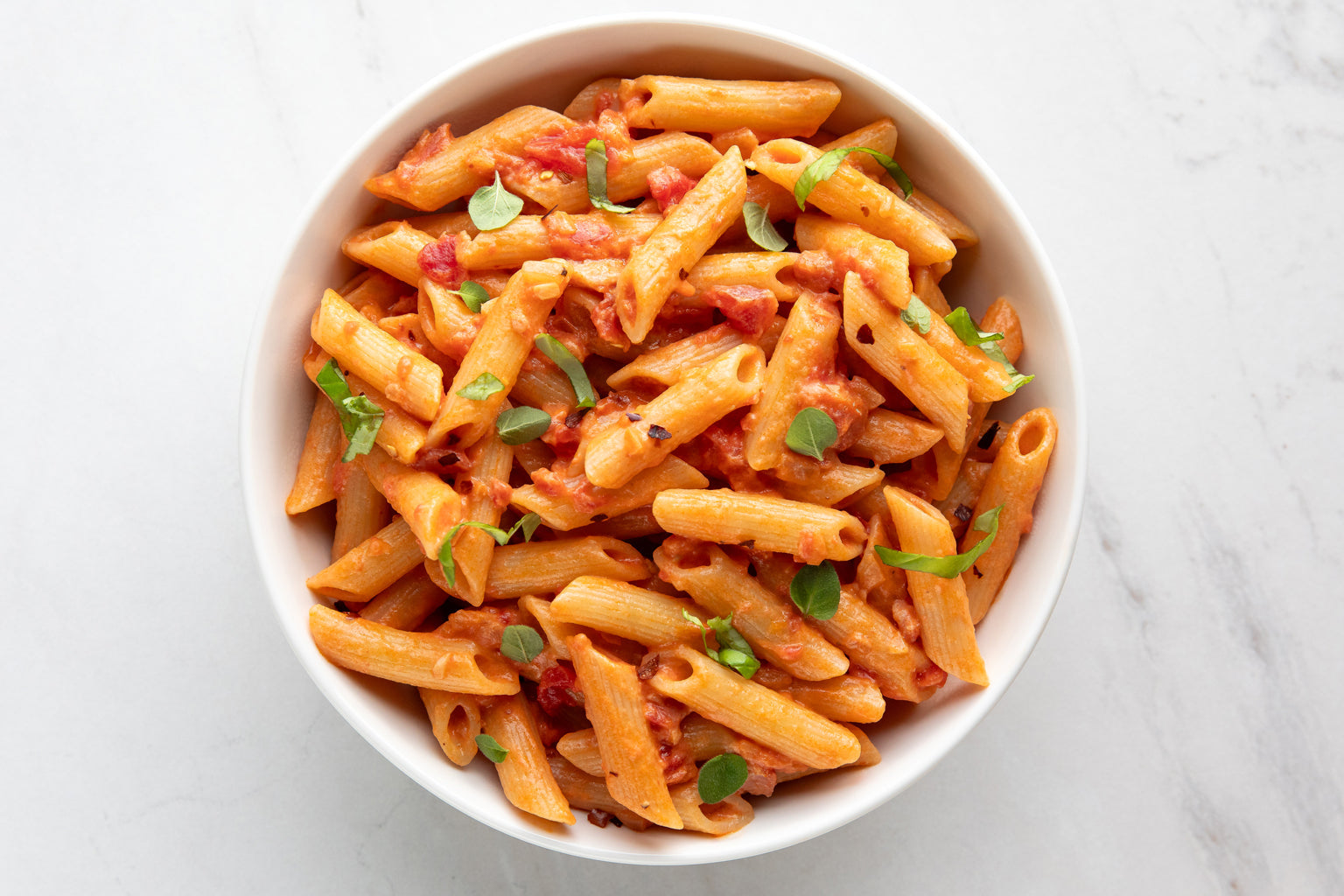 Bowl of penne pasta in creamy tomato vodka sauce garnished with fresh basil on a white marble surface