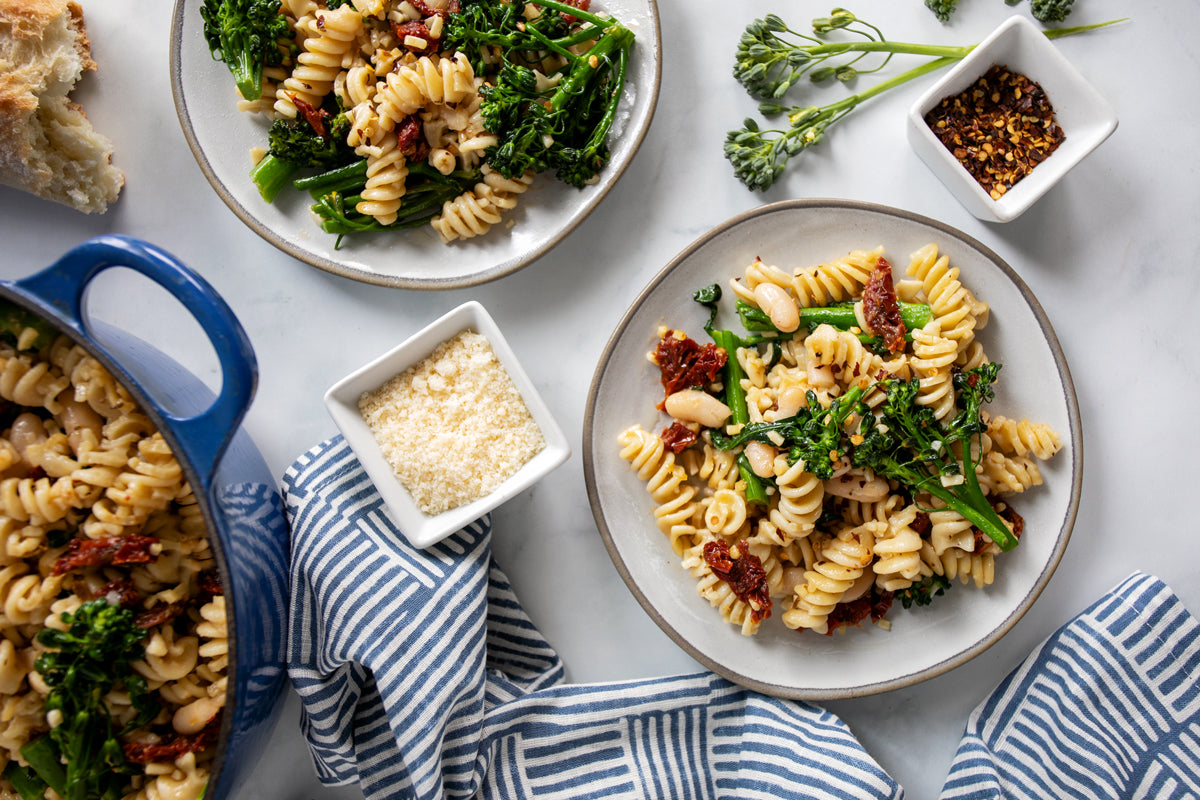 Two plates of fusilli with rapini and cannellini beans, sun-dried tomatoes, and grated cheese on a marble table