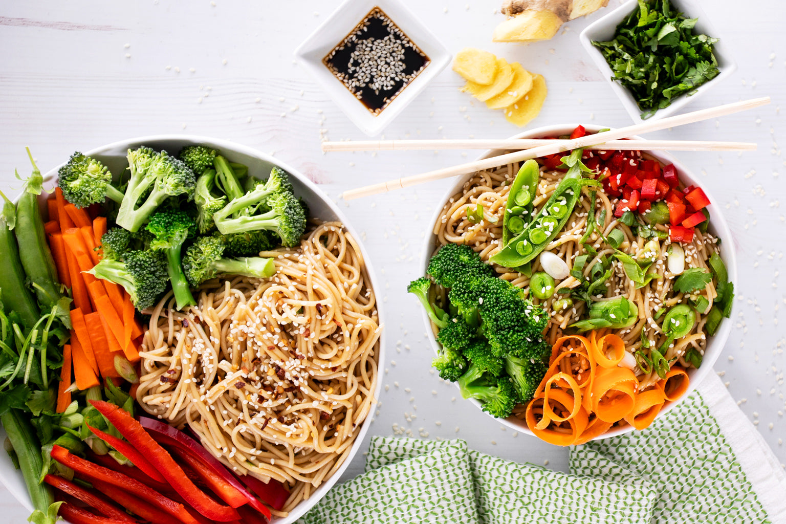 Two bowls of sesame scallion ginger noodles with broccoli, carrot ribbons, snap peas, and red pepper on a white surface