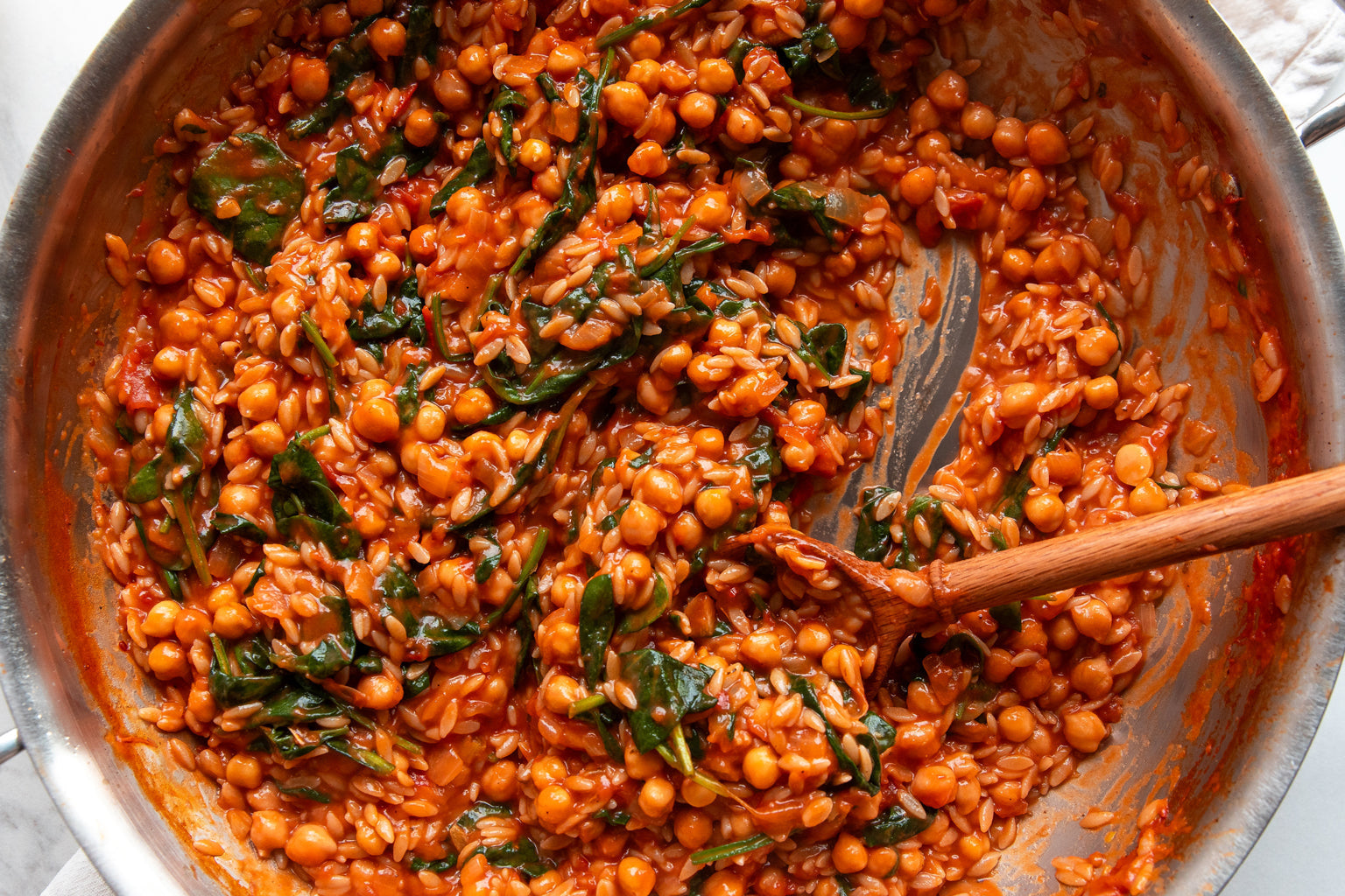 Pan of saucy tomato chickpeas and orzo being stirred with a wooden spoon, wilted greens visible