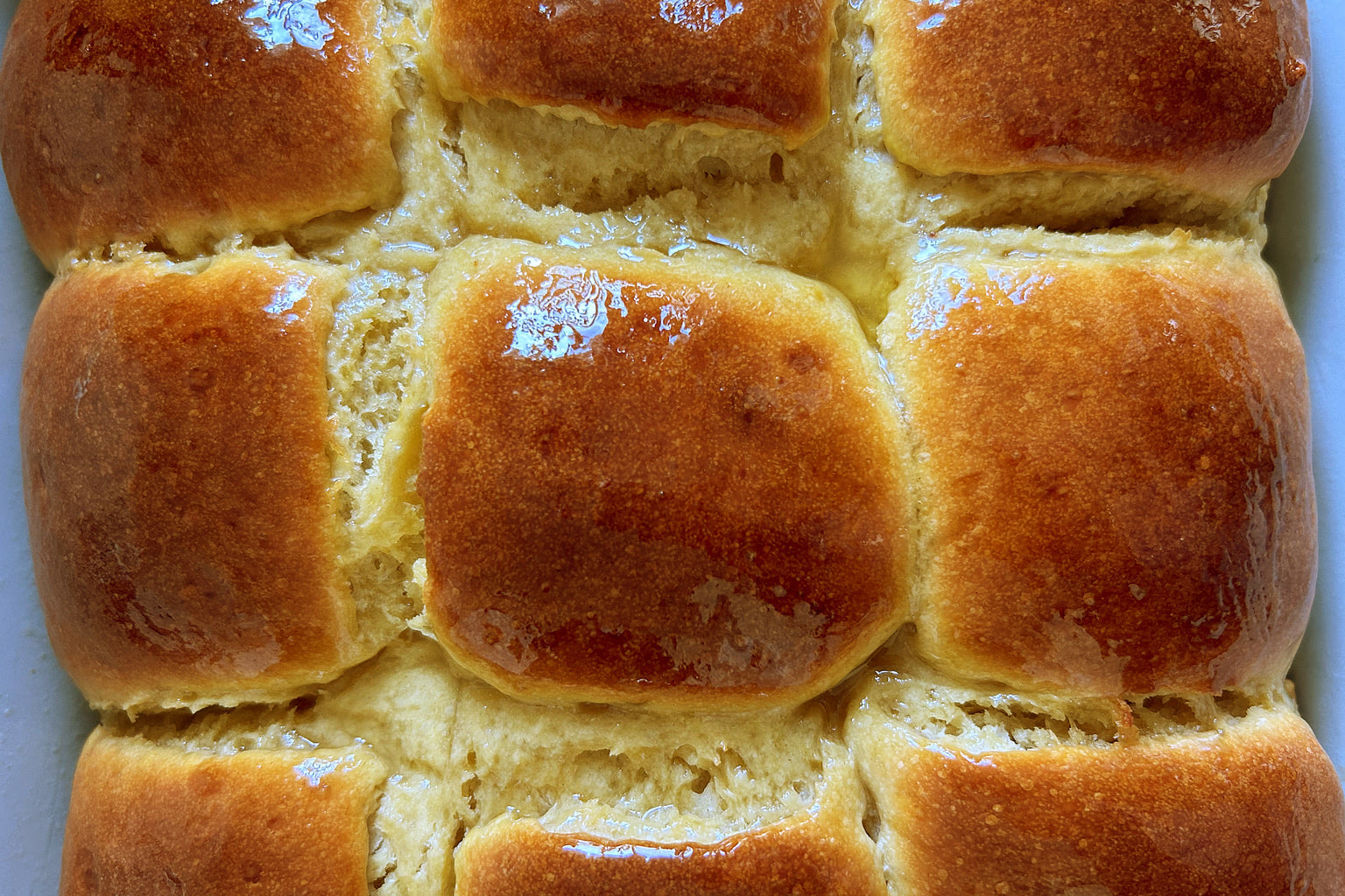 Overhead close-up of nine sourdough sweet Hawaiian rolls with glossy golden-brown tops in a white baking dish