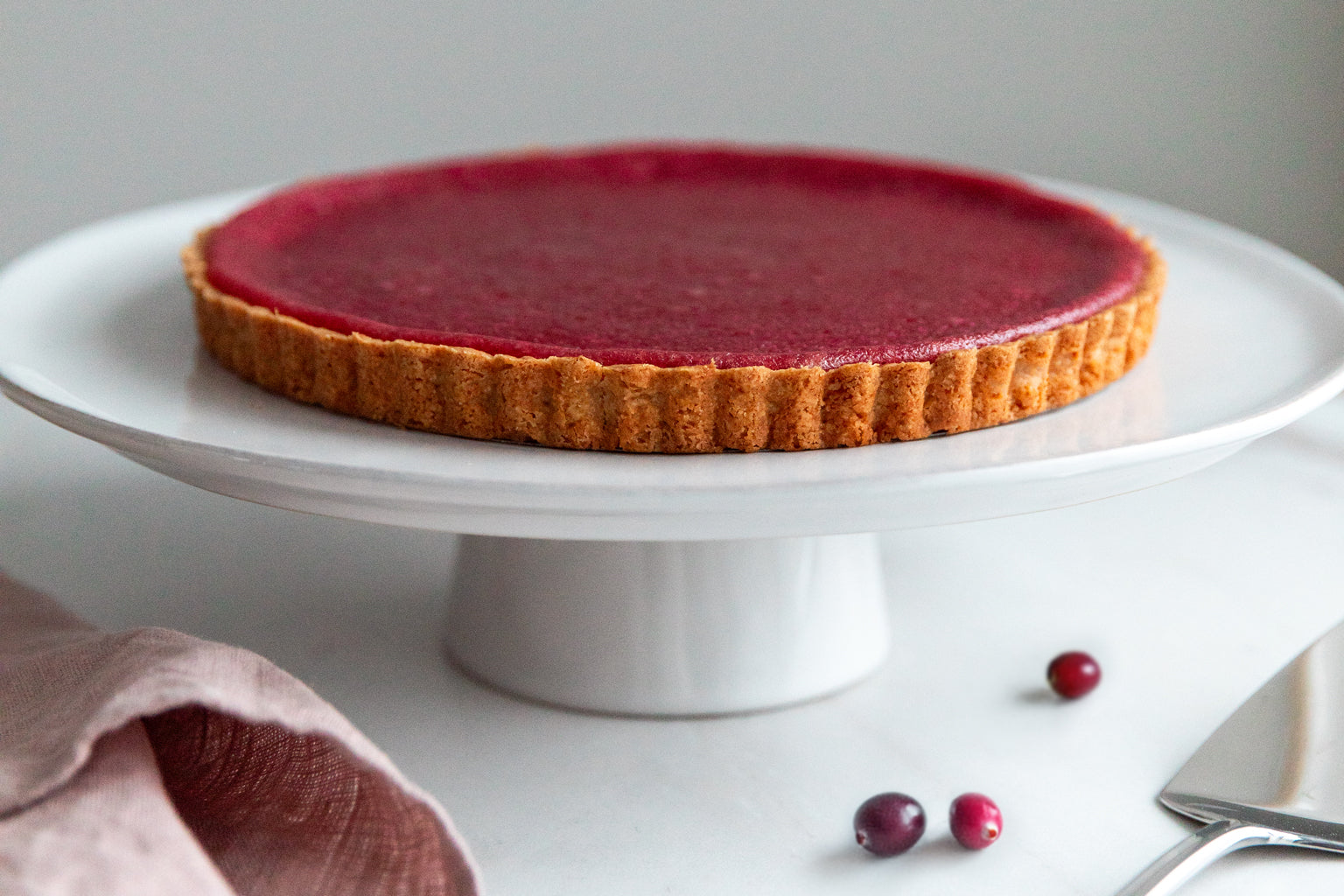 Side view of an Einkorn cranberry tart on a white cake stand showing glossy cranberry filling and fluted golden crust