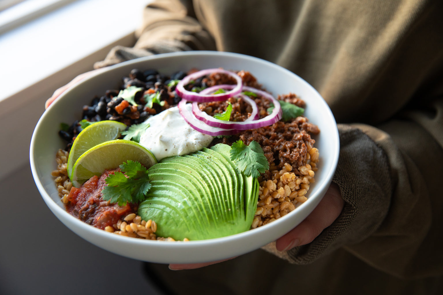 Einkorn wheat berry taco bowl with sliced avocado, black beans, salsa, lime wedges, sour cream, seasoned crumbles, red onion rings, and cilantro held near a window