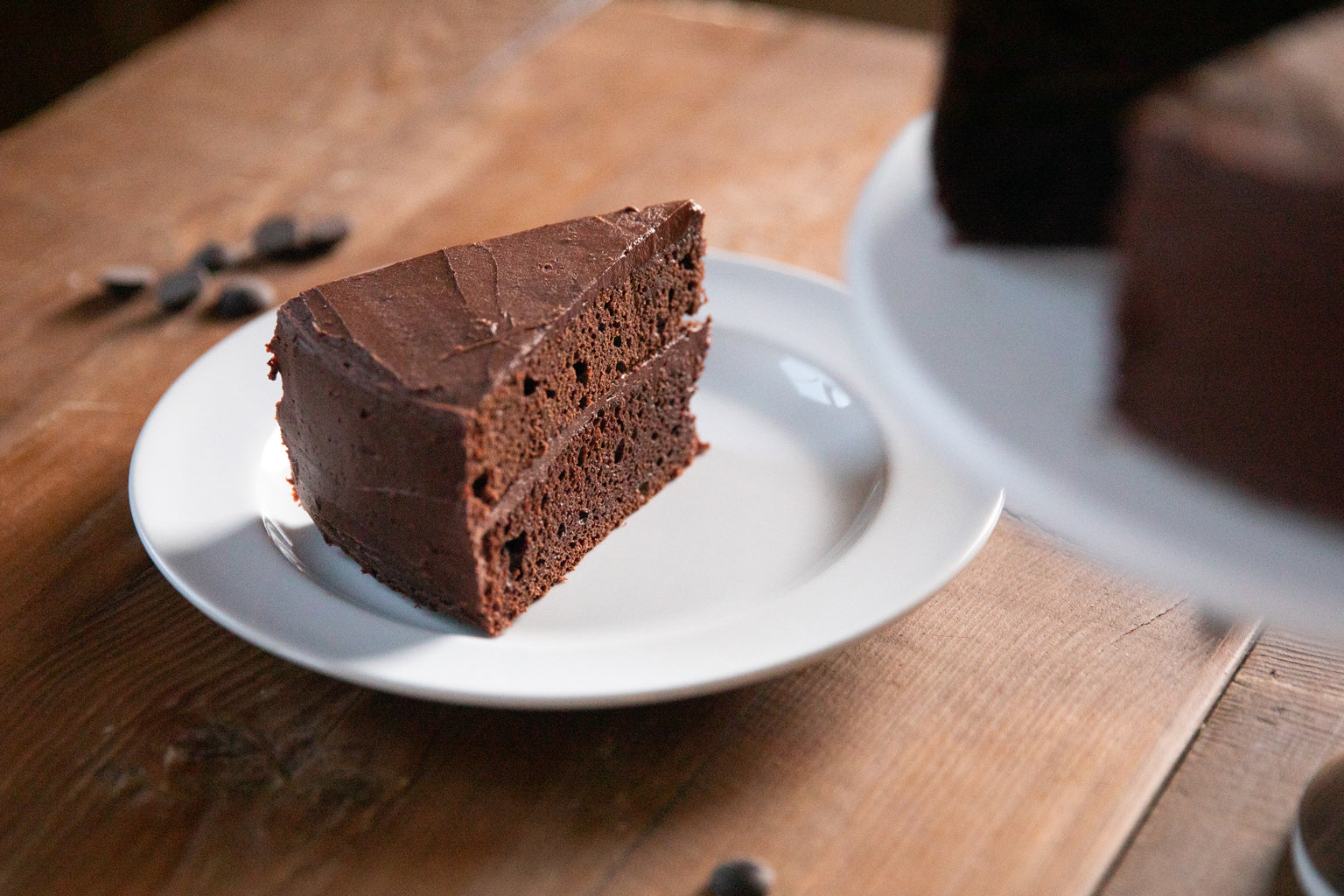 Side view of a slice of fudgy mocha Einkorn cake with two layers and chocolate frosting on a white plate on a wooden table