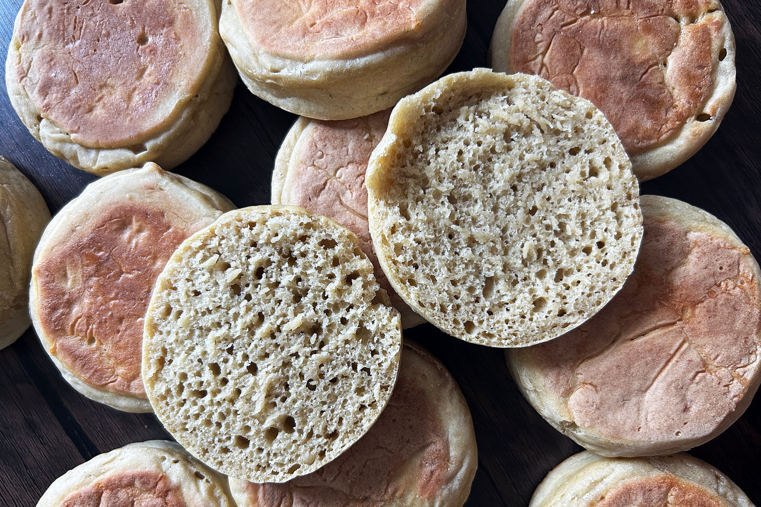 Overhead view of sourdough Einkorn English muffins, one split to reveal airy crumb and golden-brown griddle tops on dark board