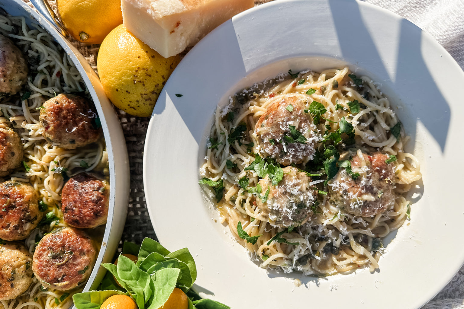 Chicken piccata meatballs on capellini in a white bowl topped with grated Parmesan and parsley, lemons and a skillet beside