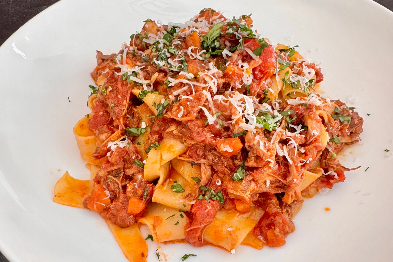 Overhead of pappardelle with pork and short rib ragù on a white plate, pasta with chunky tomato meat sauce and grated cheese