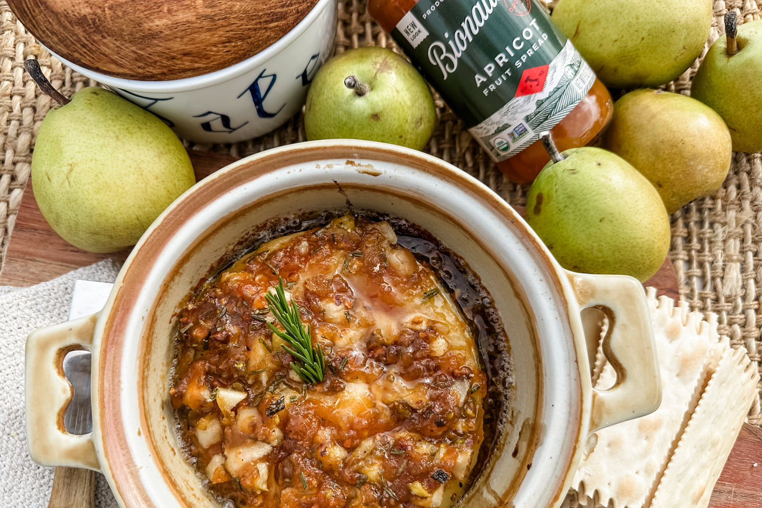 Baked brie topped with apricot spread and rosemary in a ceramic ramekin, served with crackers, pears, and a jar of Bionaturae