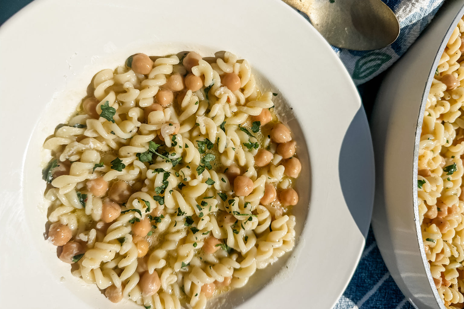 Top-down view of fusilli pasta and chickpeas tossed with olive oil and chopped parsley on a white plate beside a pan