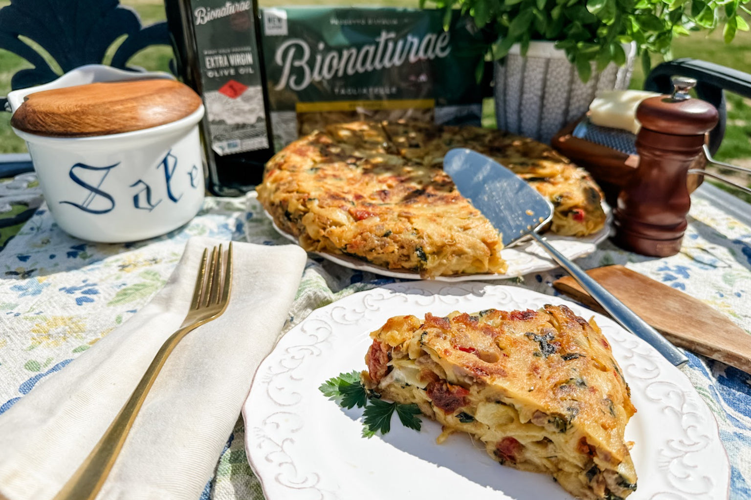 Sausage and spinach frittata slice on a white plate with the whole frittata and a Bionaturae pasta box on an outdoor table