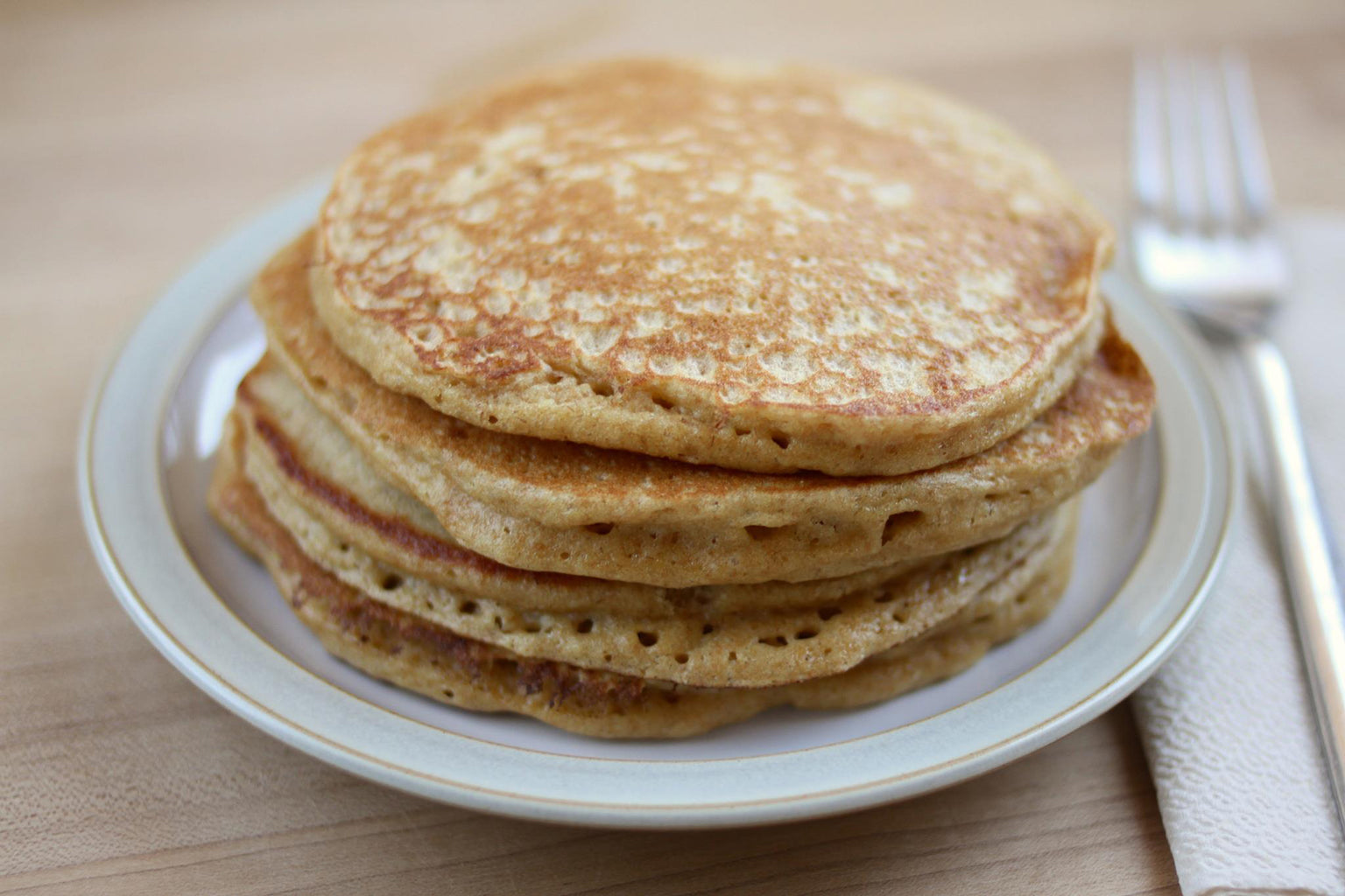 Stack of whole-grain Einkorn pancakes on a white plate with a fork and napkin on a wooden table