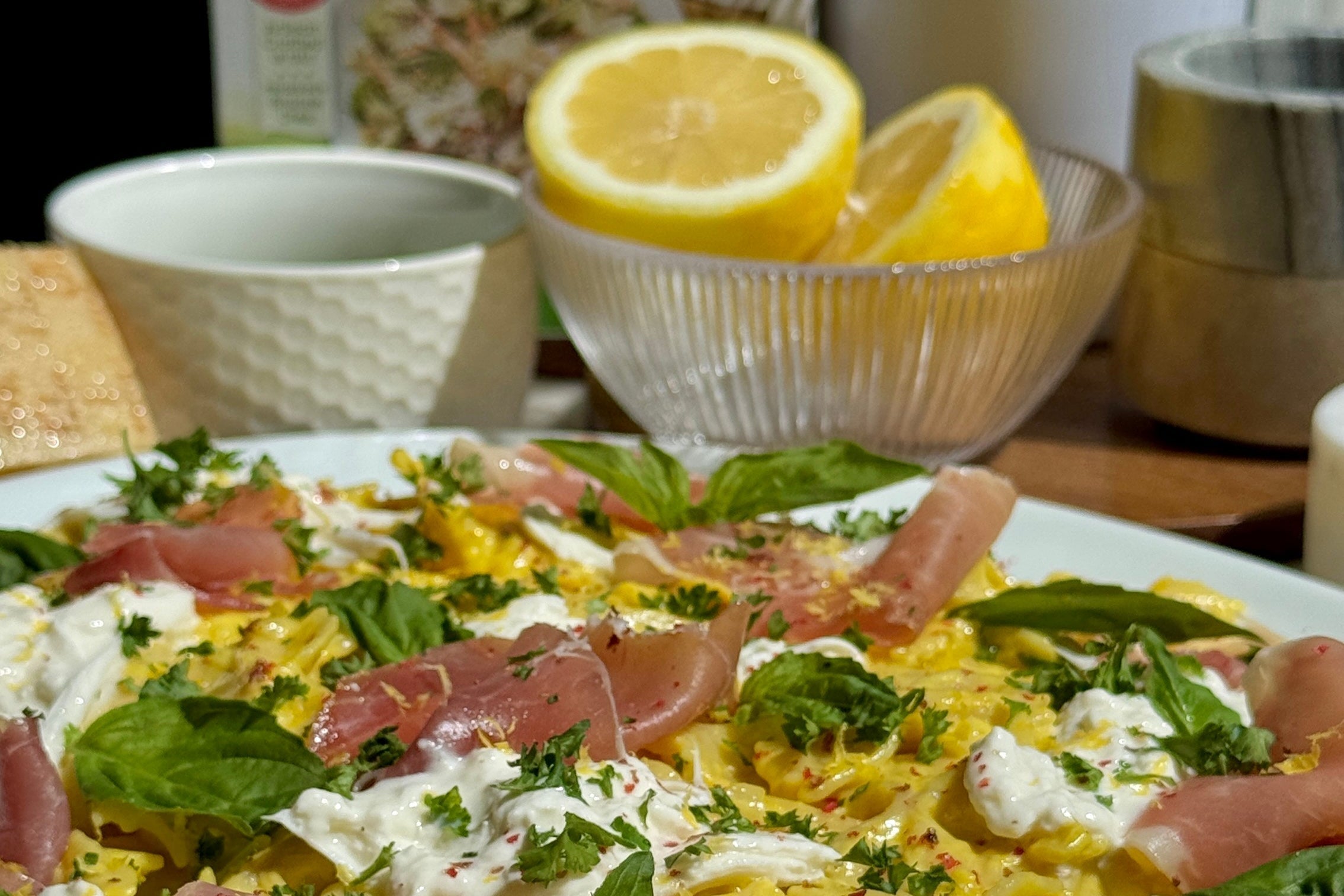 Plate of farfalle in creamy lemon-saffron sauce topped with burrata, prosciutto, basil and parsley, lemon halves behind