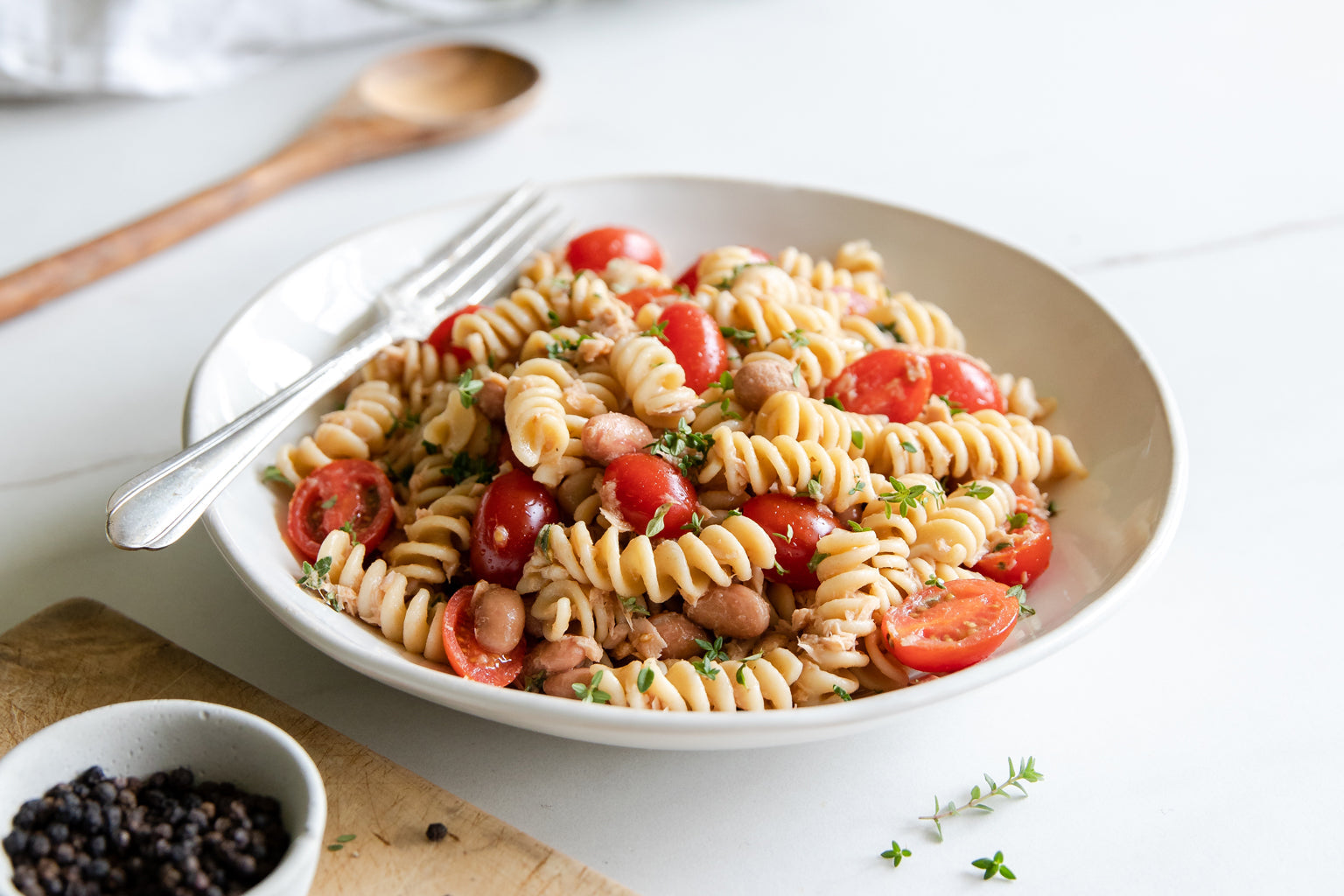 Borlotti bean and tuna rotini pasta salad with cherry tomatoes and fresh herbs in a white bowl, fork and wooden spoon nearby