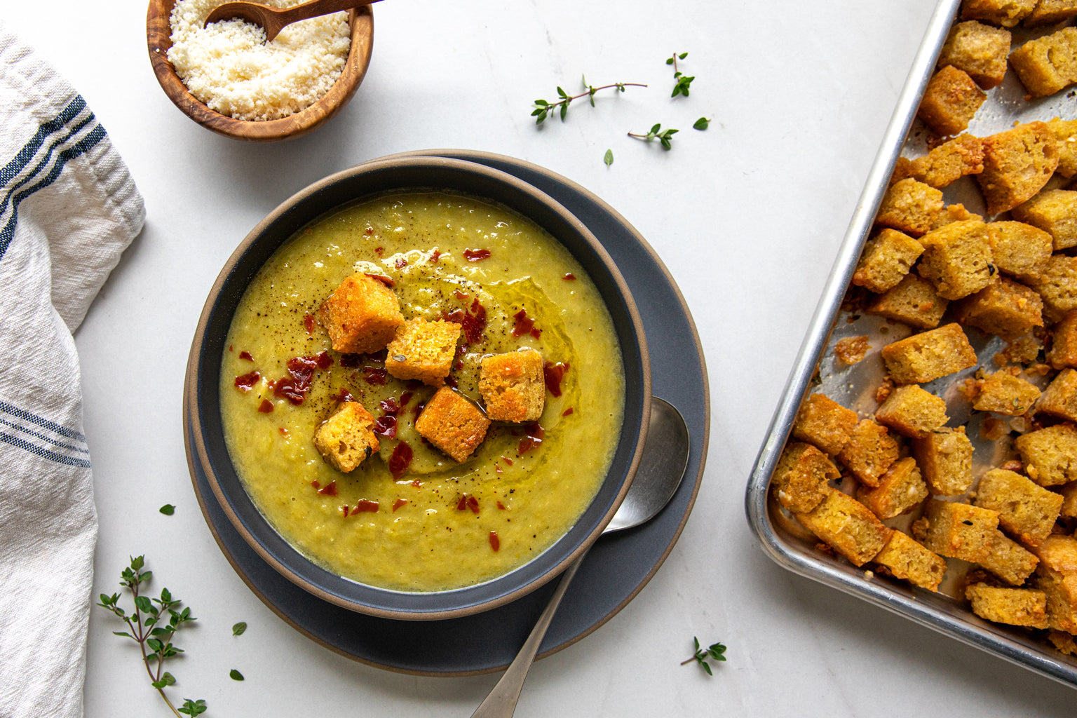 Bowl of asparagus soup topped with herby einkorn croutons beside a tray of croutons and a bowl of grated cheese