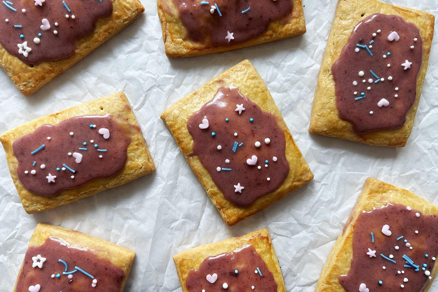 Overhead view of sourdough wild berry pop tarts with purple berry glaze and sprinkles on crumpled parchment paper