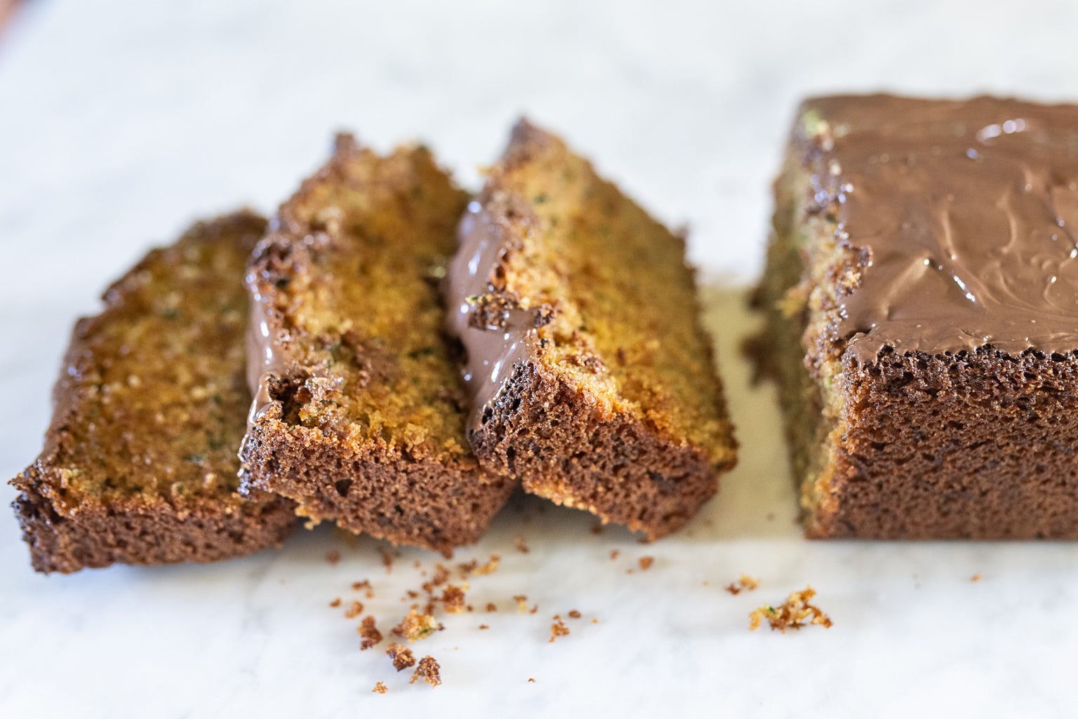 Close-up of sliced rustic zucchini bread with chocolate-hazelnut glaze and crumbly texture on a white marble surface