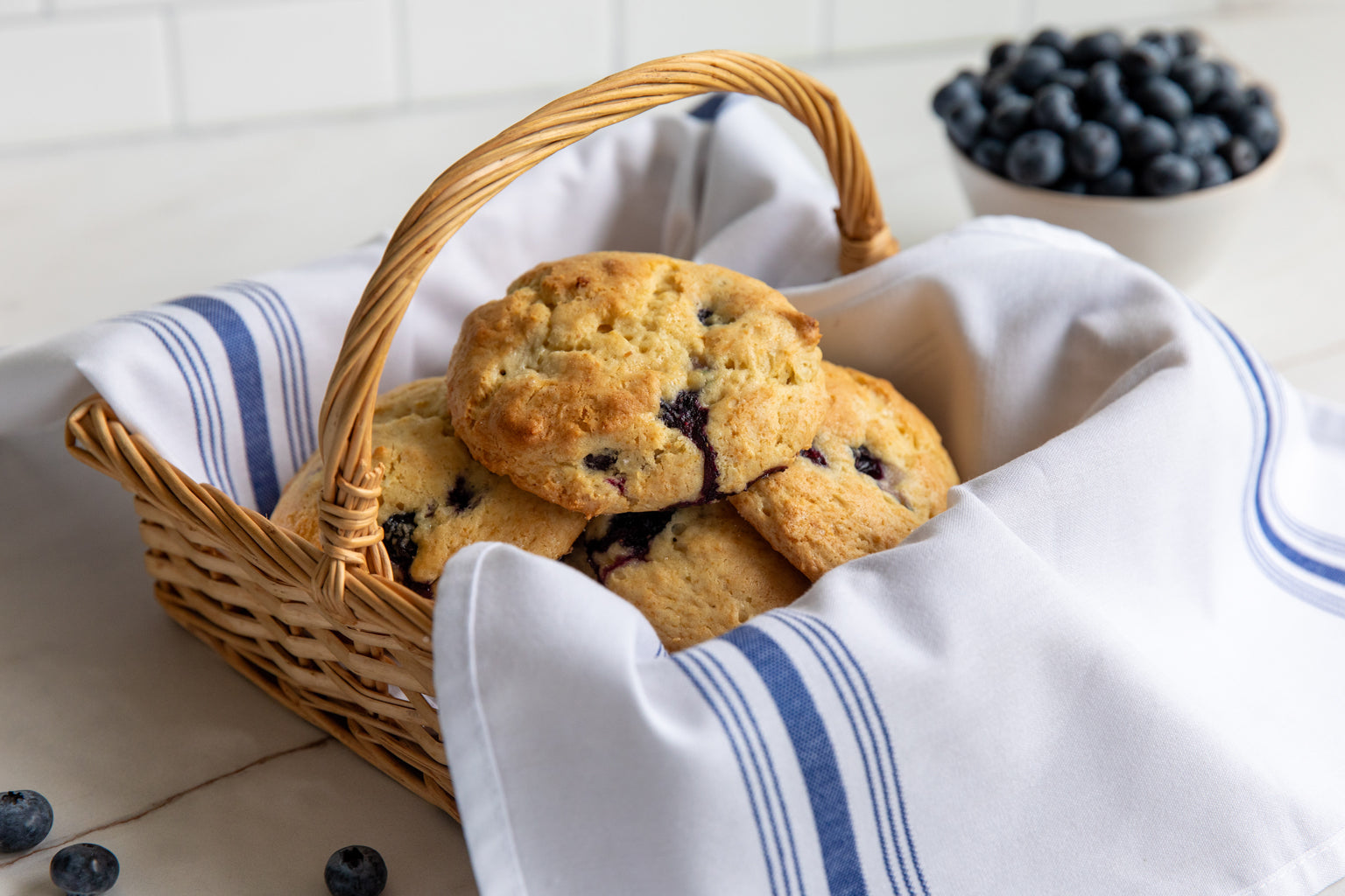 Einkorn blueberry drop scones in a wicker basket on a blue-striped cloth with a bowl of blueberries behind